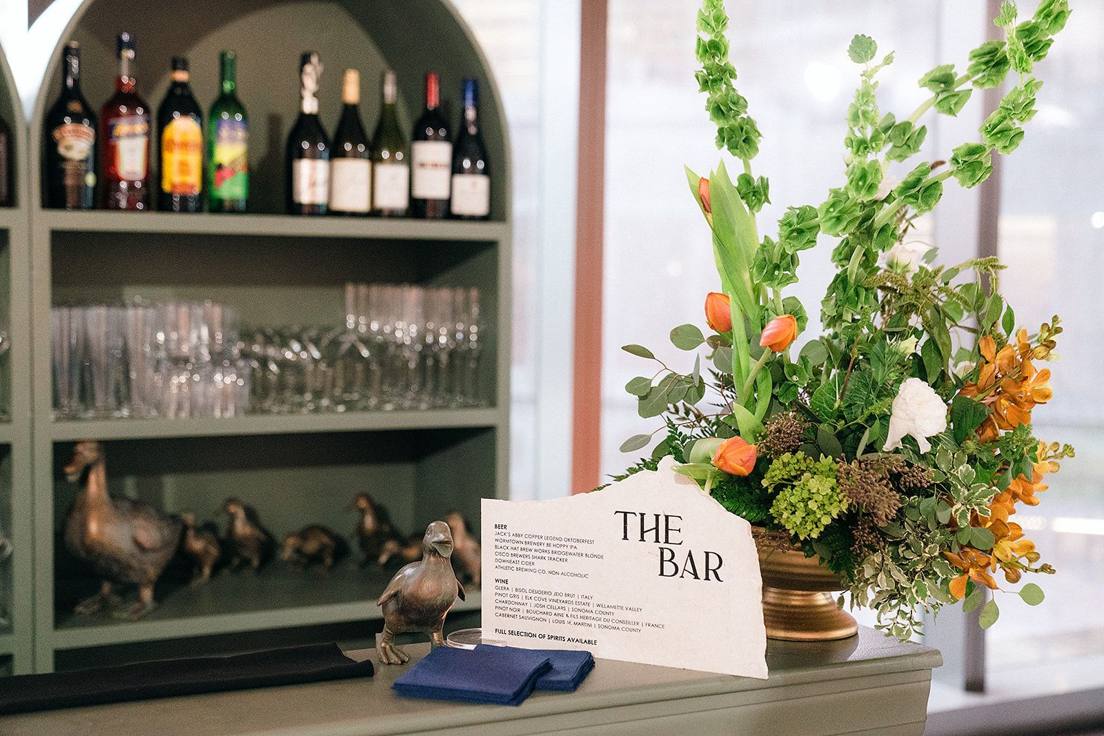 Bar area with bottles, glasses, and a floral arrangement; a menu sign reads 