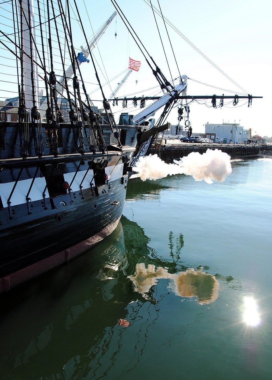 Tall black ship with masts and rigging firing a cannon over calm harbor water