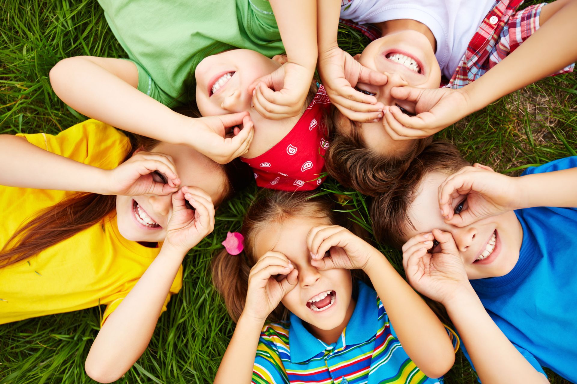 Six smiling children lie in a circle on green grass, making heart shapes with their hands.
