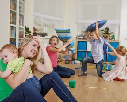 A woman is holding a baby while a group of children are playing with pillows in a living room.