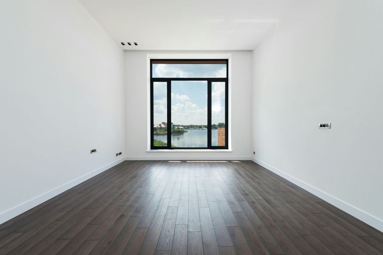 Empty room with dark wood floor, white walls, large window overlooking water.