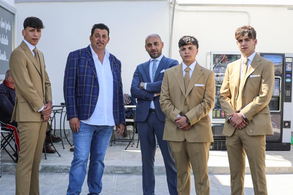 Cinque uomini fuori da un edificio, tre in abiti color sabbia e due in abiti casual, guardano la telecamera.