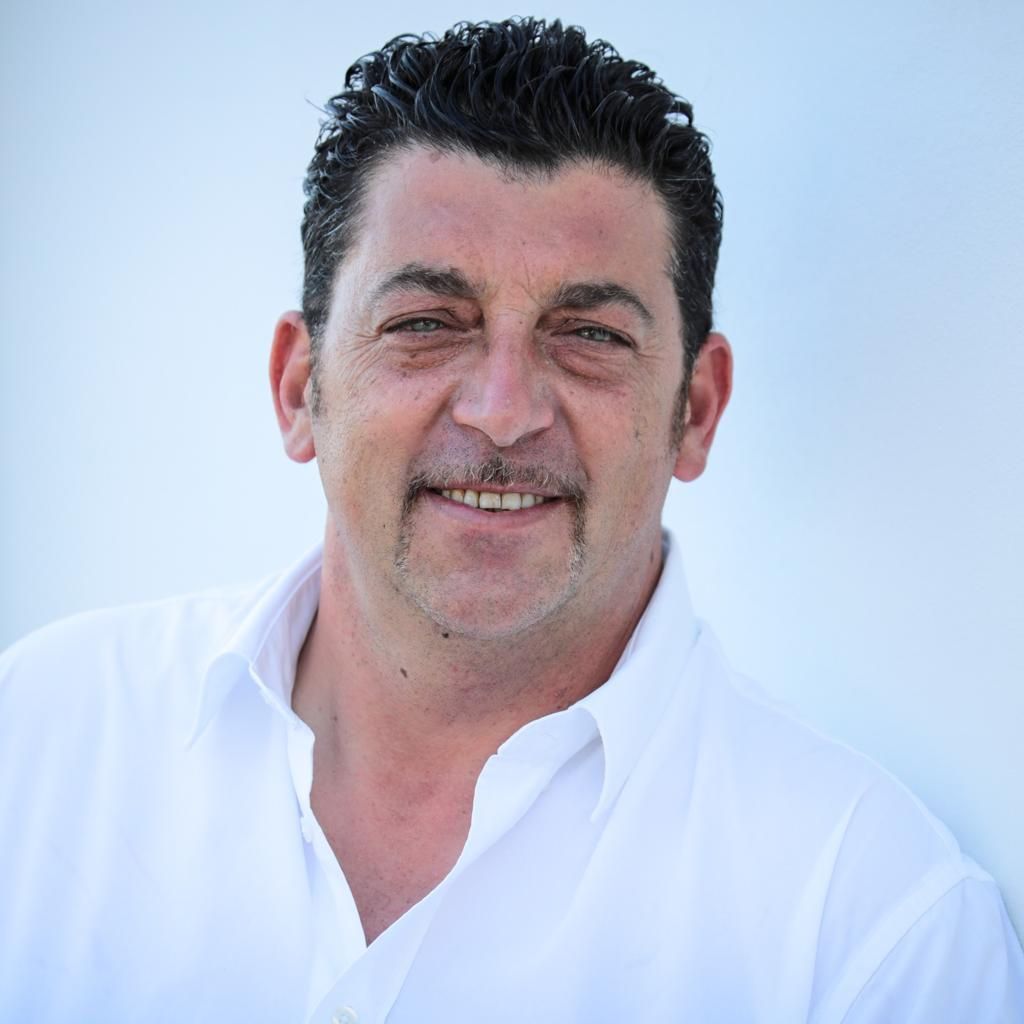 Un uomo con i capelli scuri e i baffi sorride indossando una camicia bianca su uno sfondo bianco.