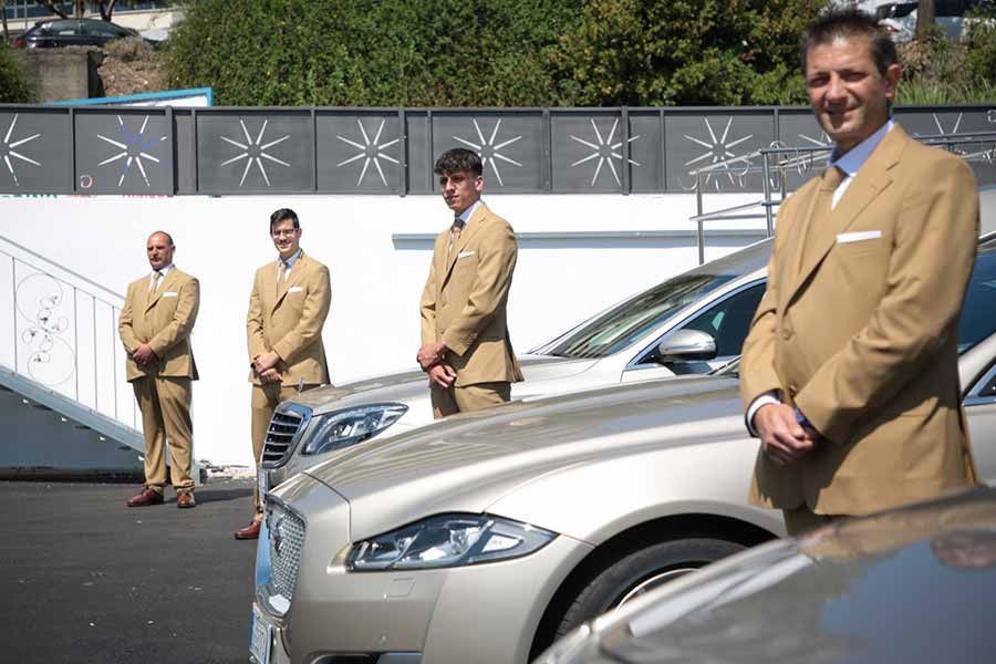 Quattro uomini vestiti in modo formale in piedi vicino ad auto di lusso; abiti color sabbia