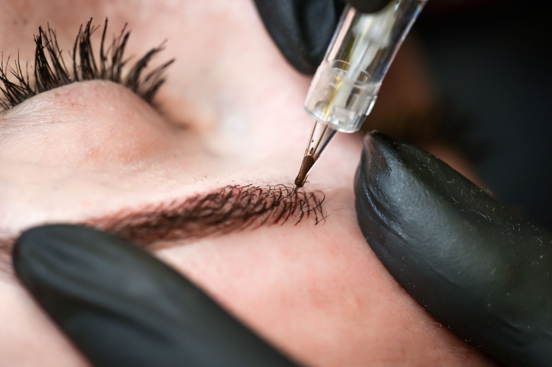 Close-up of gloved hands tattooing an eyebrow with a fine needle.