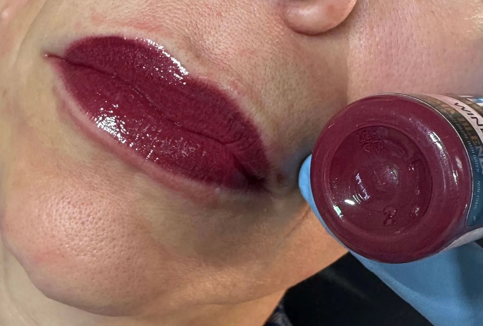 Close-up of glossy burgundy lips while a gloved hand holds a matching lip gloss wand nearby