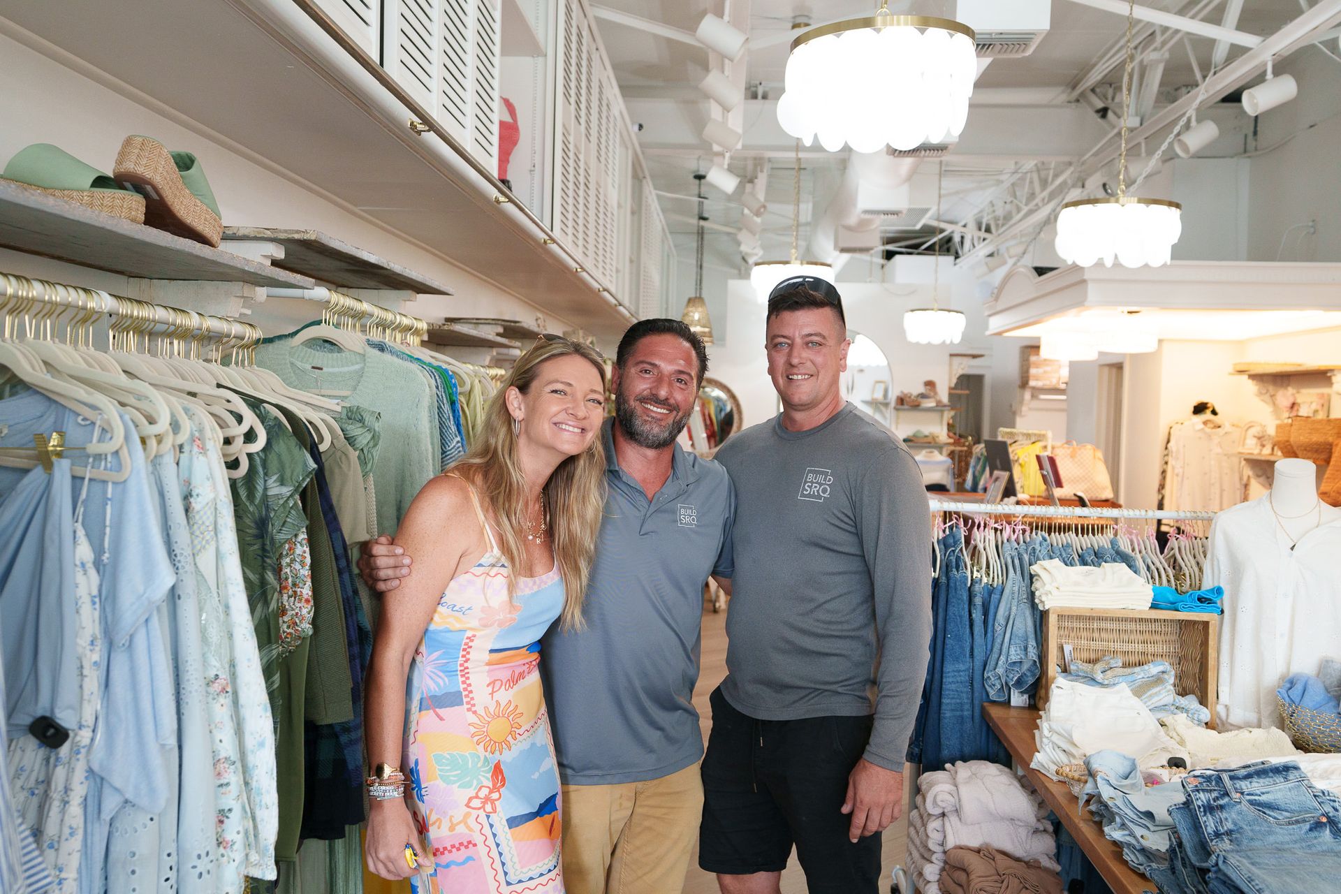 Three smiling people standing in a bright clothing boutique among racks of dresses and decor