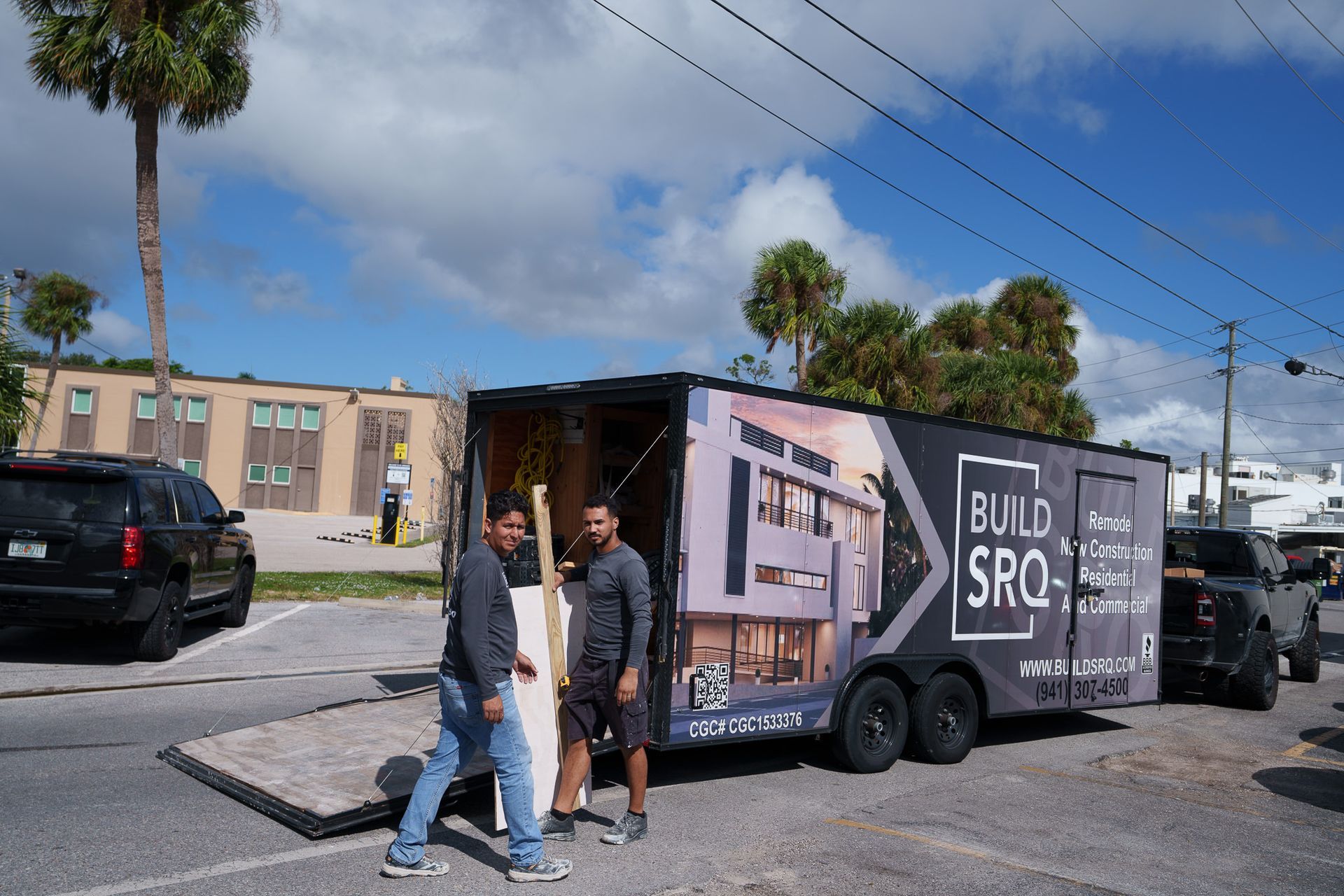 Two people walking past a black trailer with large “BUILD, SRO” graphics on a sunny street.