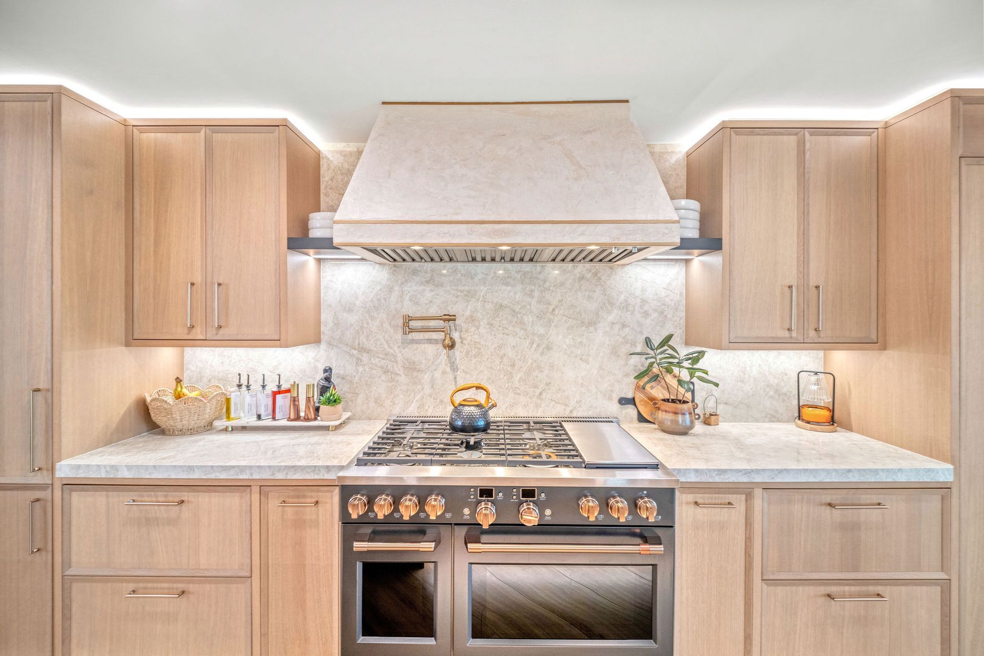 Bright modern kitchen with light wood cabinets, marble countertops, and a stainless steel range with hood.