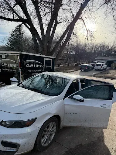 White sedan with open driver door parked on a street beside a leafless tree and utility trailer.