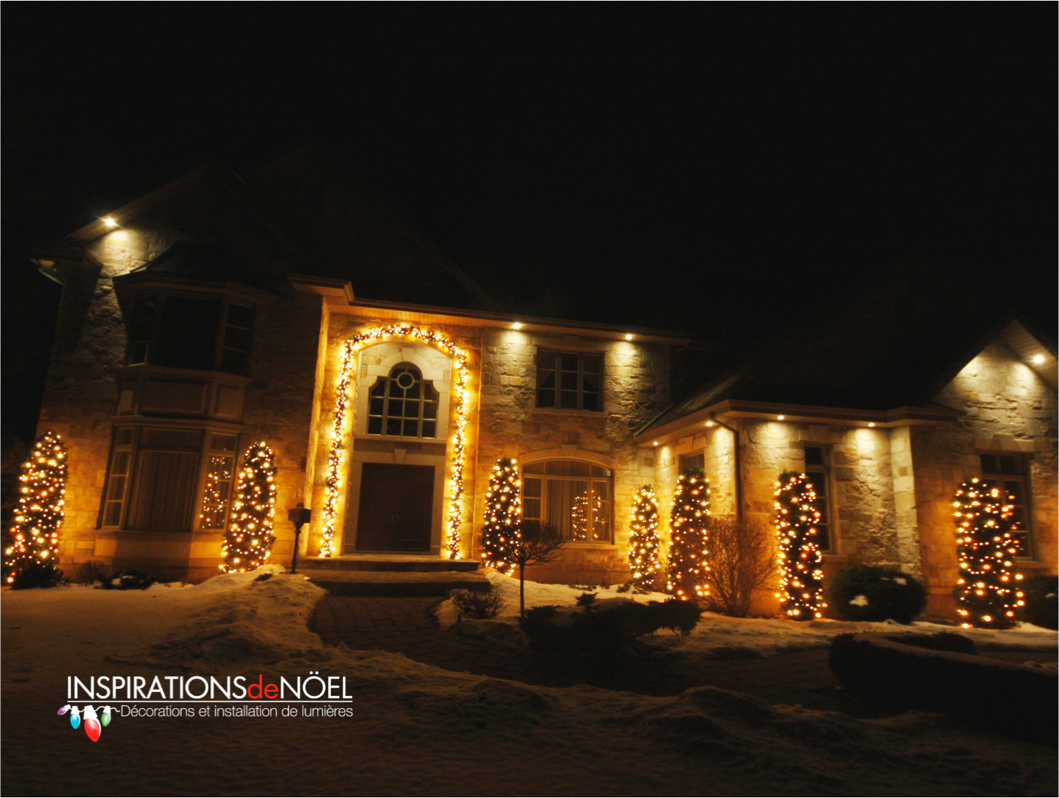 Grande maison en pierre illuminée de lumières de Noël la nuit, neige au sol.