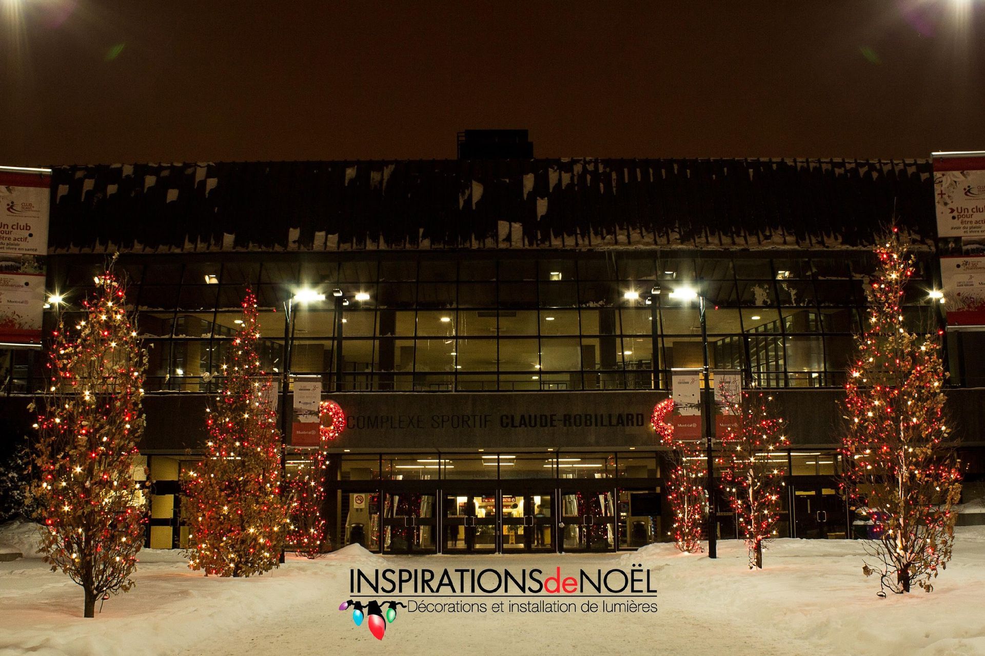 Entrée du bâtiment la nuit, sapins de Noël illuminés, sol enneigé.
