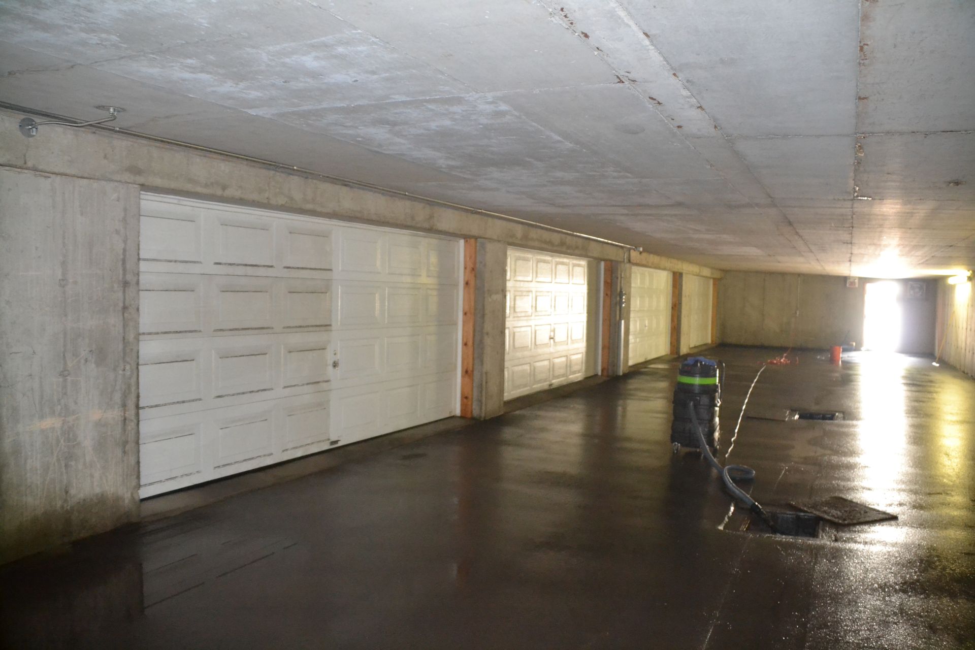 Parking souterrain avec plusieurs portes blanches et sol humide et réfléchissant. Une lumière vive au bout du tunnel.