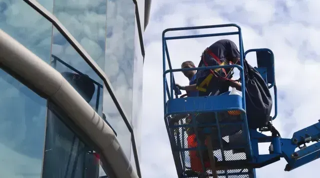 Des ouvriers dans un ascenseur bleu nettoient les vitres d'un immeuble moderne en verre réfléchissant. Journée ensoleillée.