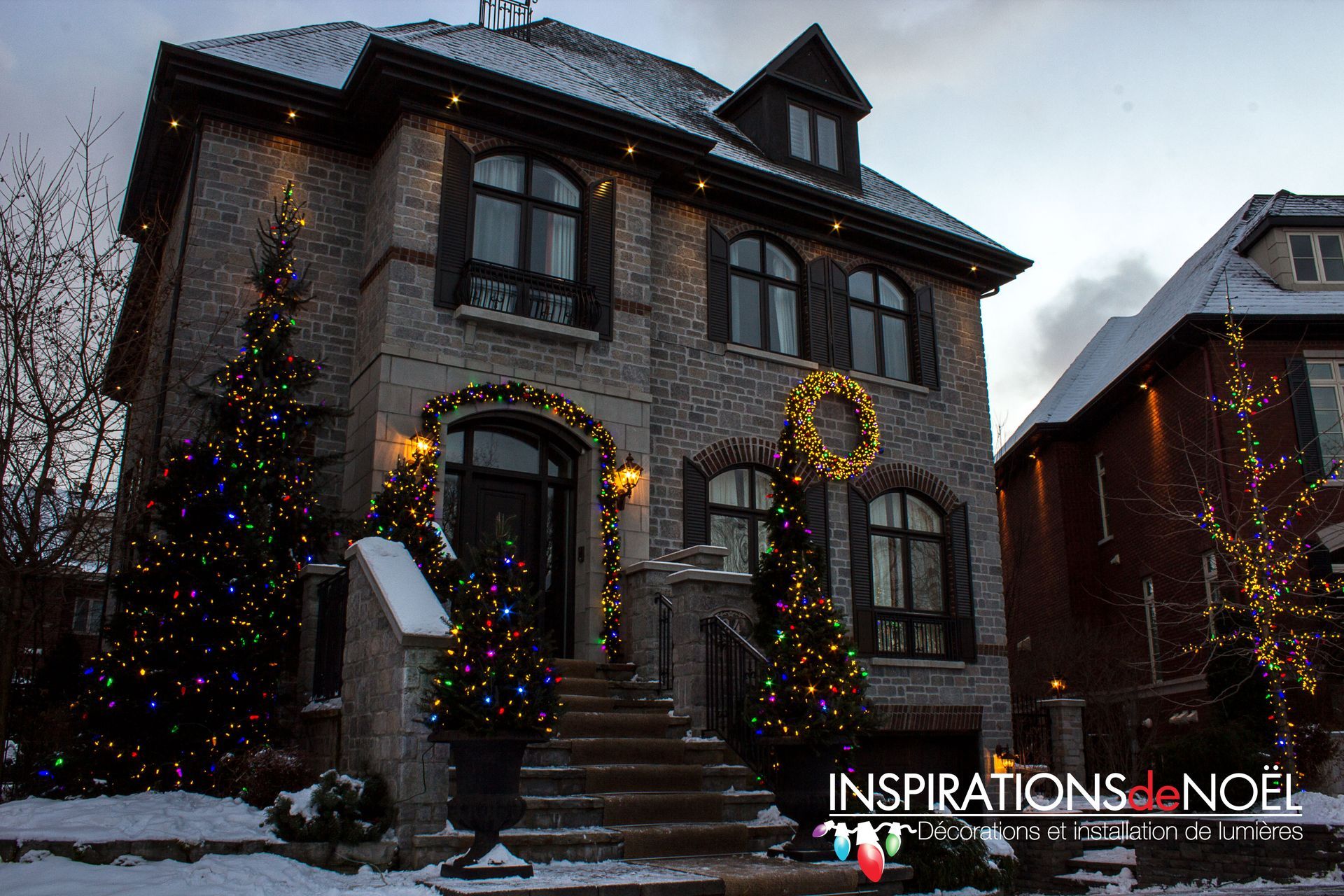Une maison en briques décorée pour Noël, avec des lumières, des guirlandes et des couronnes, placée dans la neige.