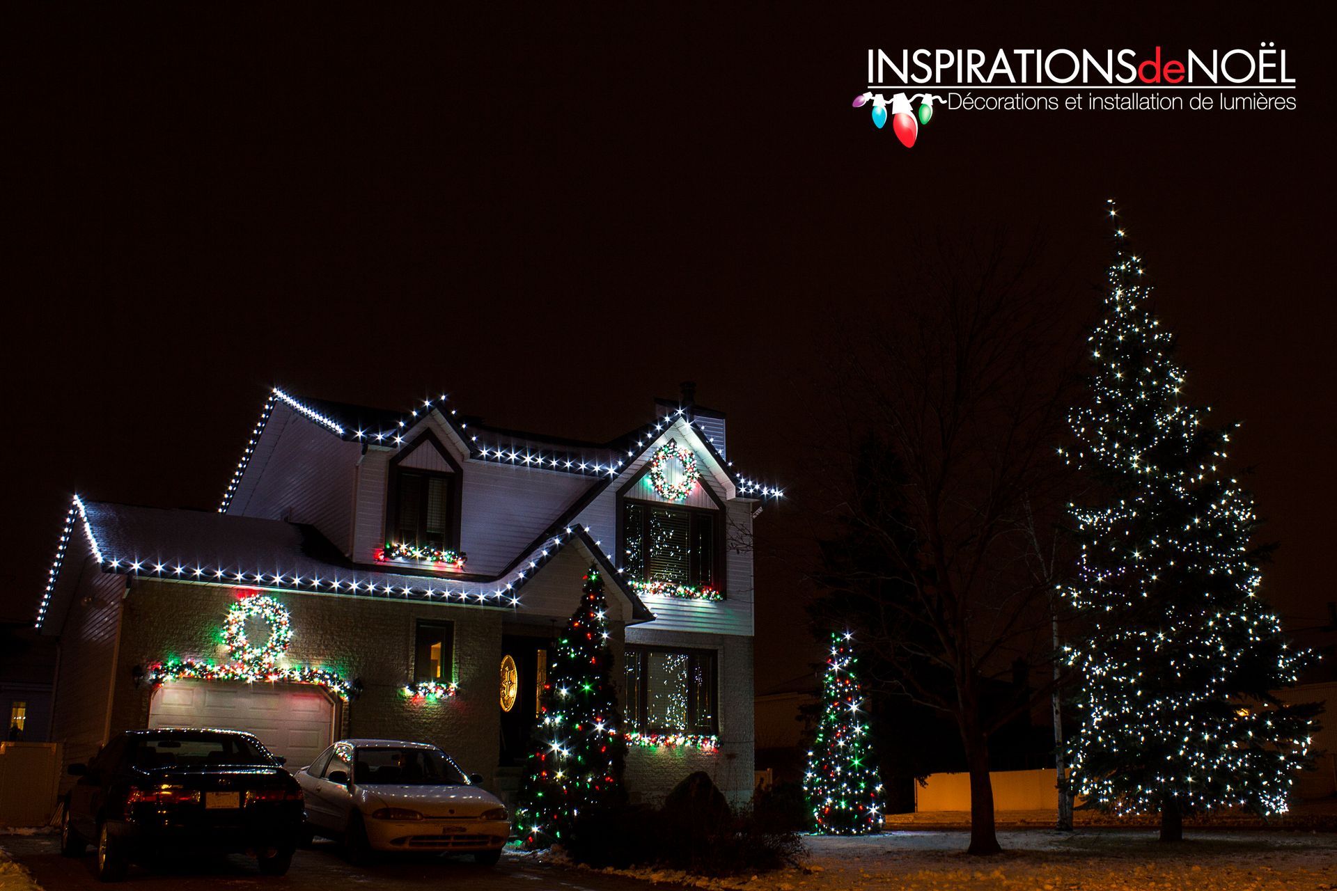 Maison décorée de lumières de Noël la nuit, avec des arbres illuminés et des voitures dans l'allée.