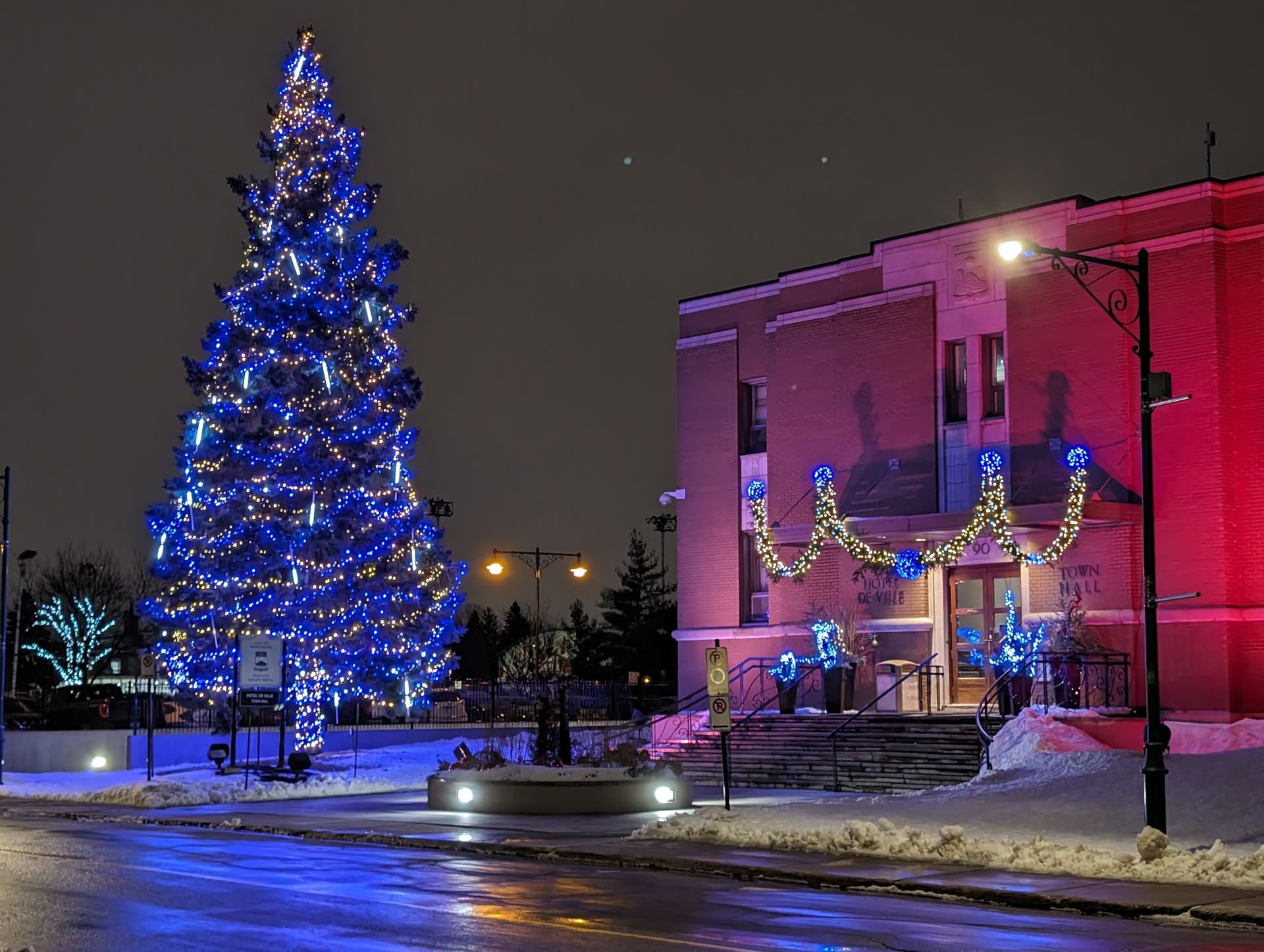 Sapin de Noël et bâtiment illuminés de lumières bleues et rouges la nuit. Neige au sol.