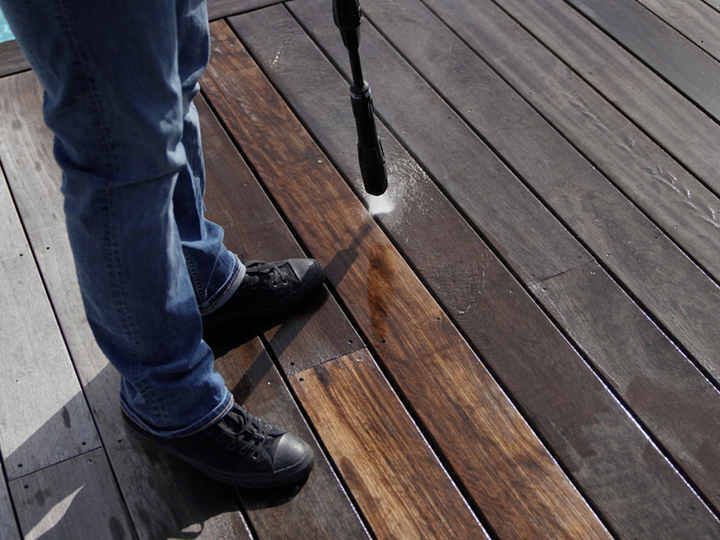 Une personne nettoie une terrasse en bois à haute pression. Le bois nettoyé paraît plus clair que le bois sale.