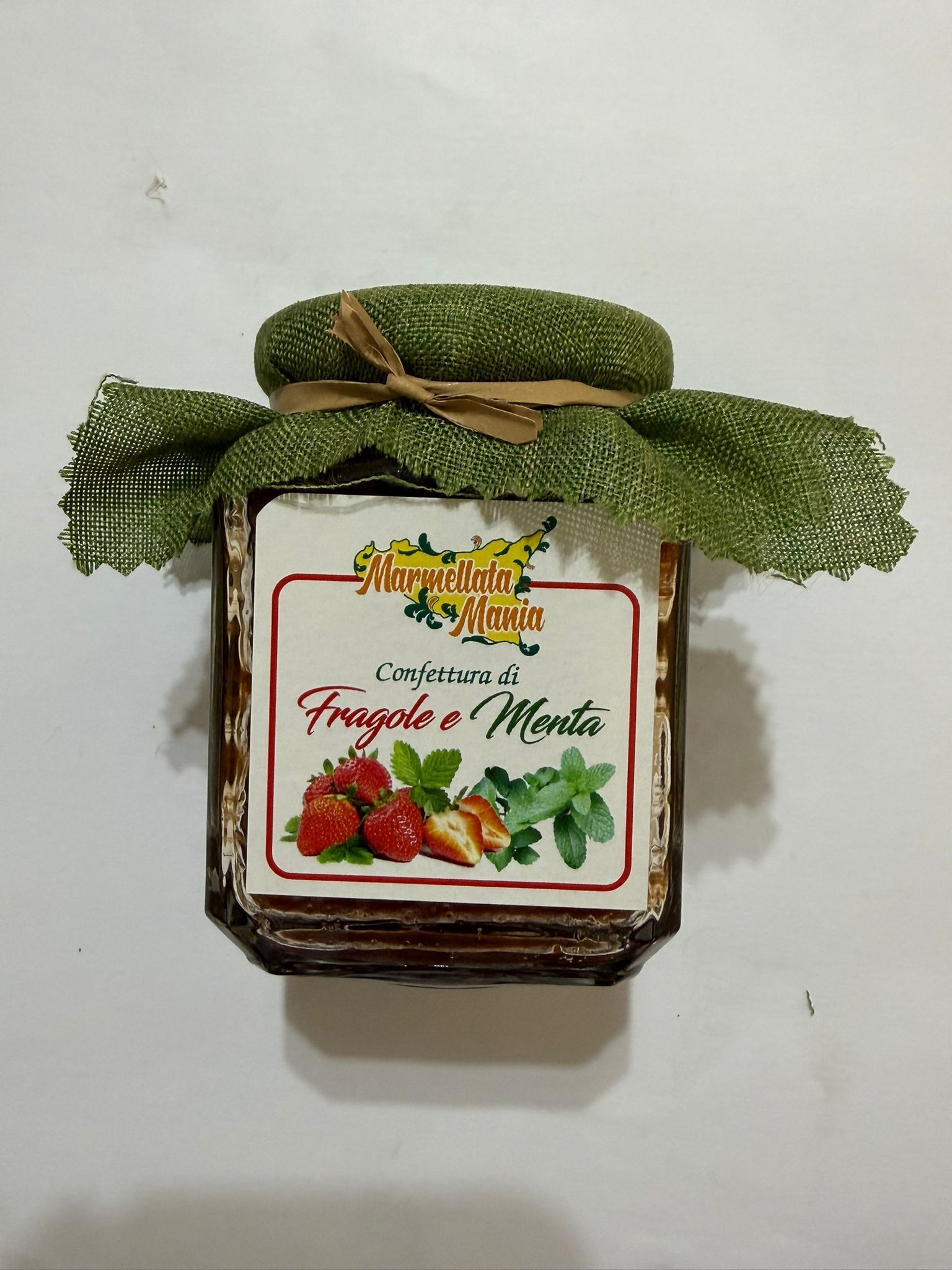 Vasetto di marmellata di fragole con coperchio in tessuto verde ed etichetta a forma di fragola su sfondo bianco.