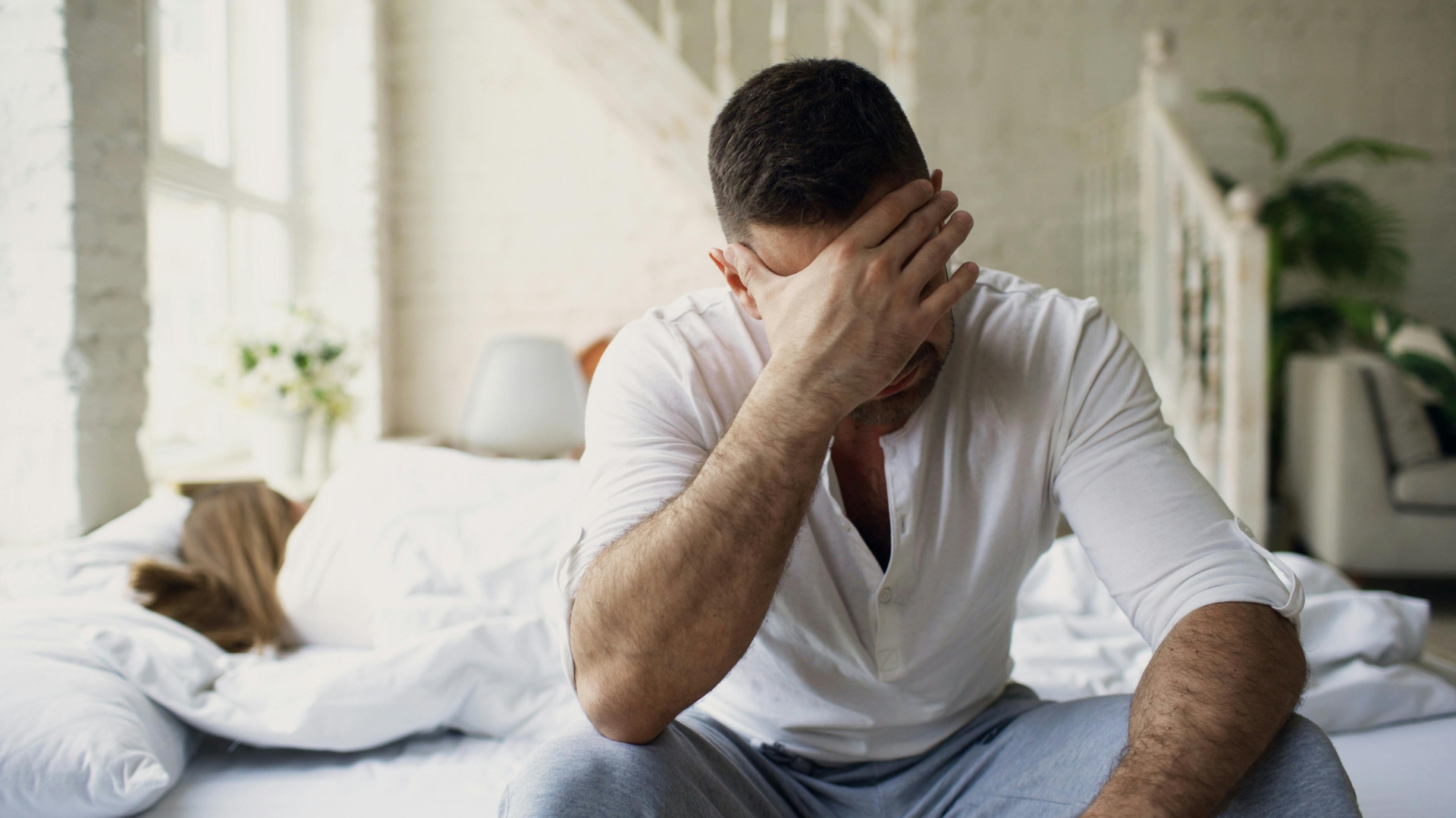 Man sitting on a bed with his hand covering his face, looking stressed in a bright bedroom.