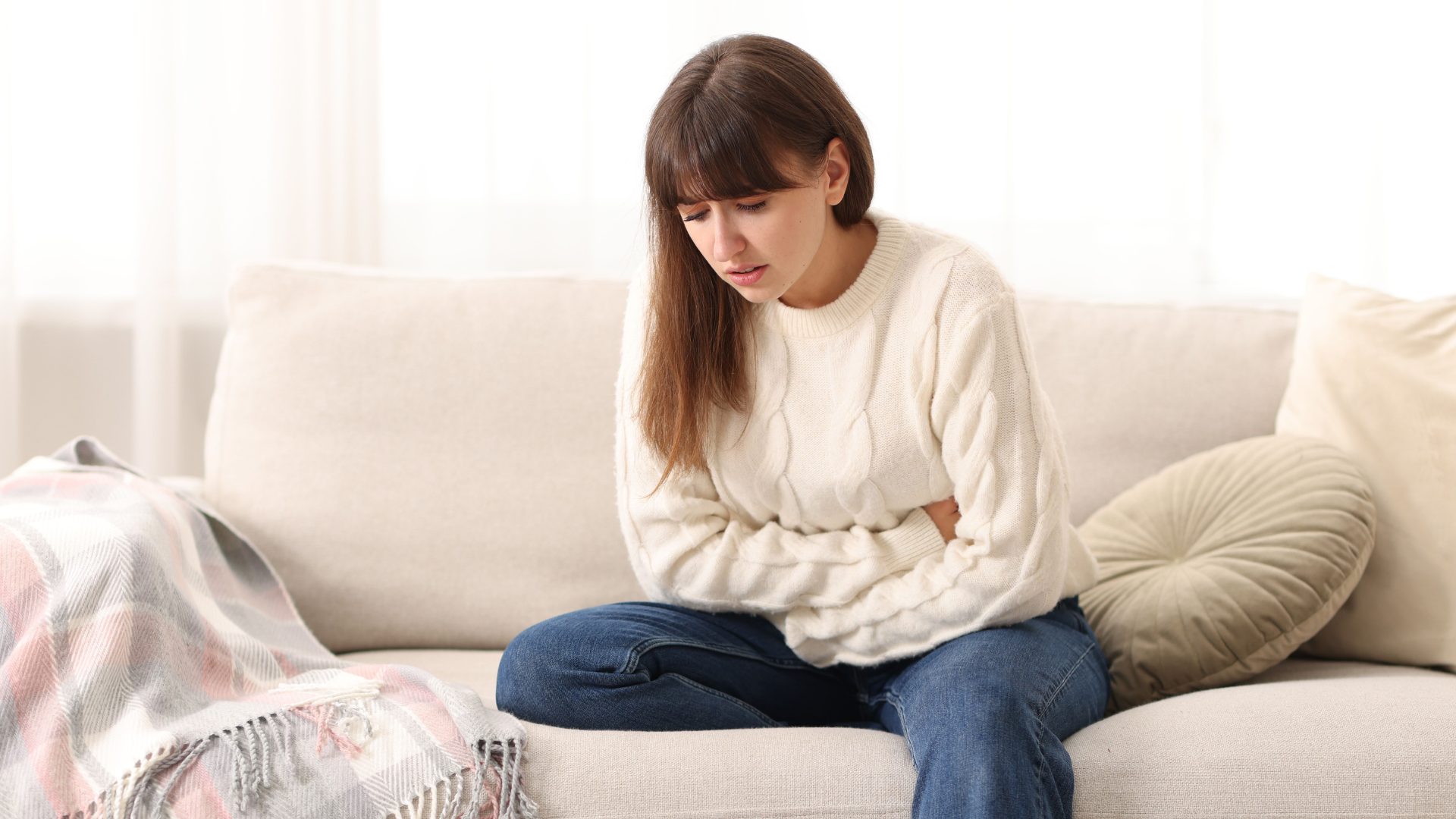 Woman sitting on a beige couch, looking down with hands clasped, appearing thoughtful or worried.