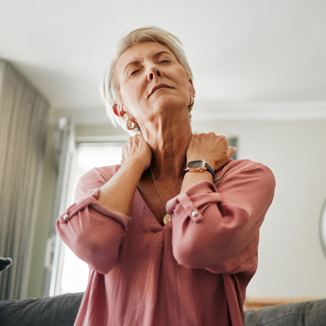 An elderly woman is sitting on a couch holding her neck in pain.