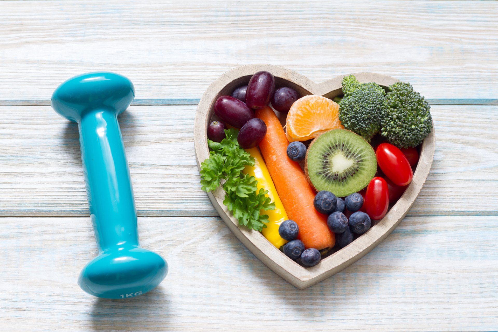 Heart-shaped fruit bowl with kiwi, berries, broccoli, lettuce, and grapes beside a blue dumbbell.