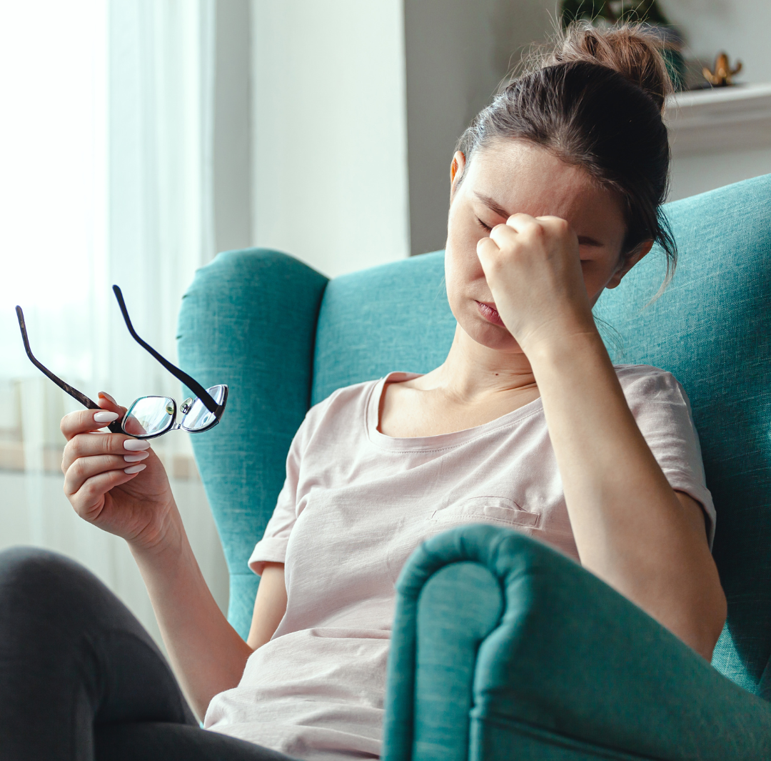 A woman is sitting in a chair holding her head and glasses.
