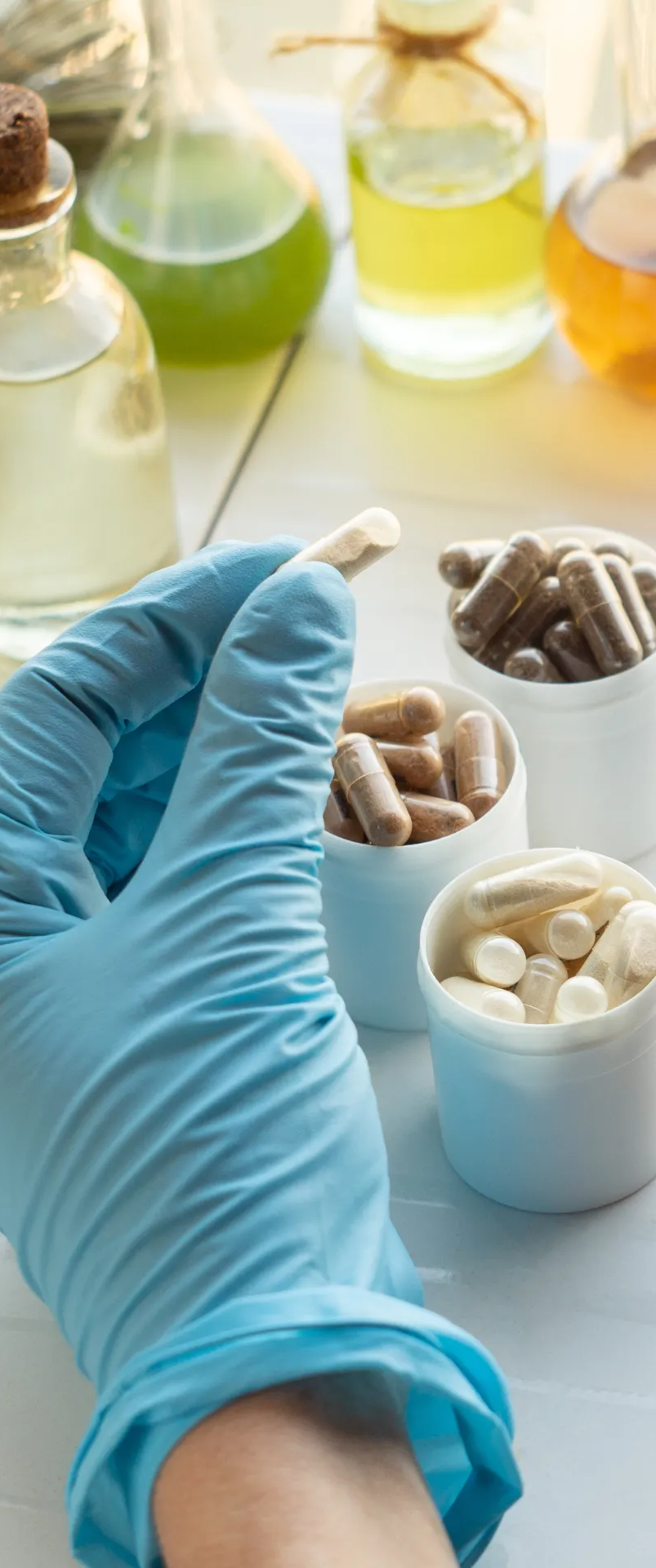 Gloved hand holding a capsule over small containers of pills in a lab setting