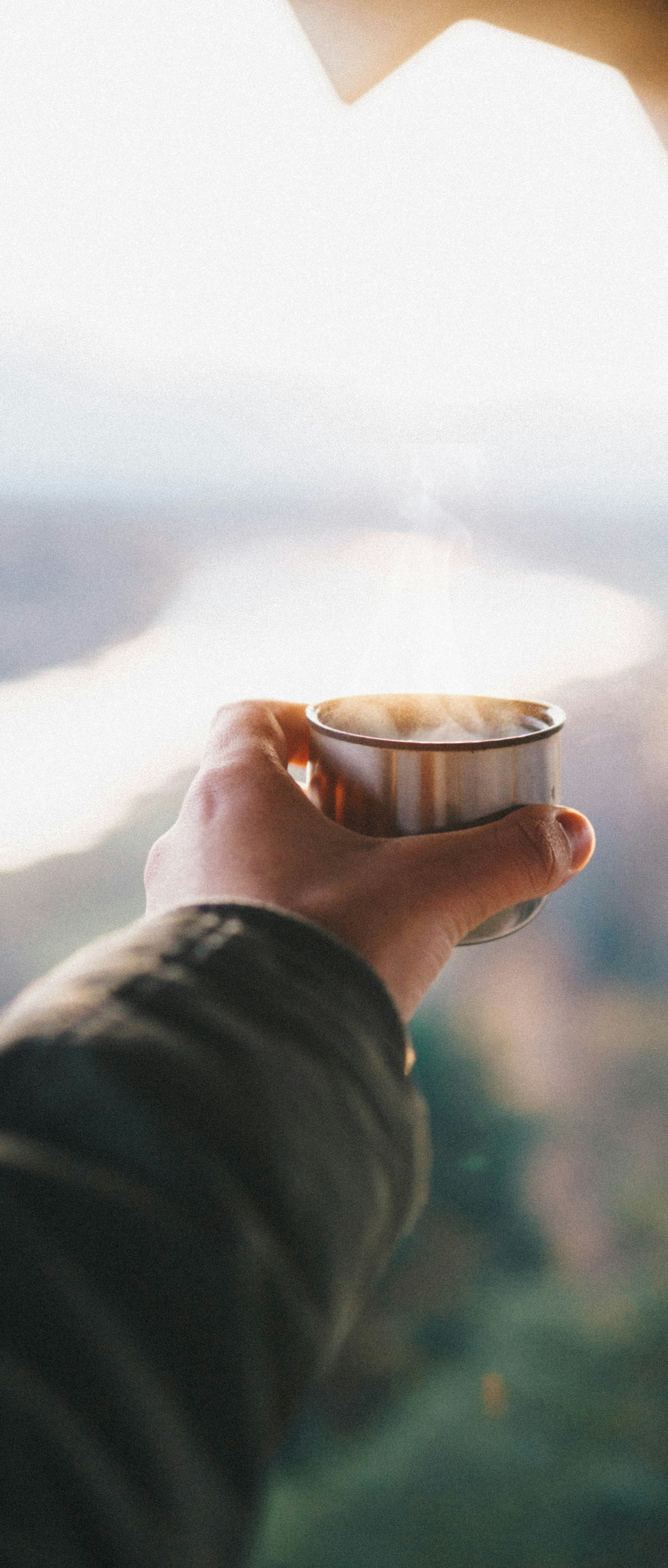 Hand holding a small cup with steam rising against a blurred outdoor background