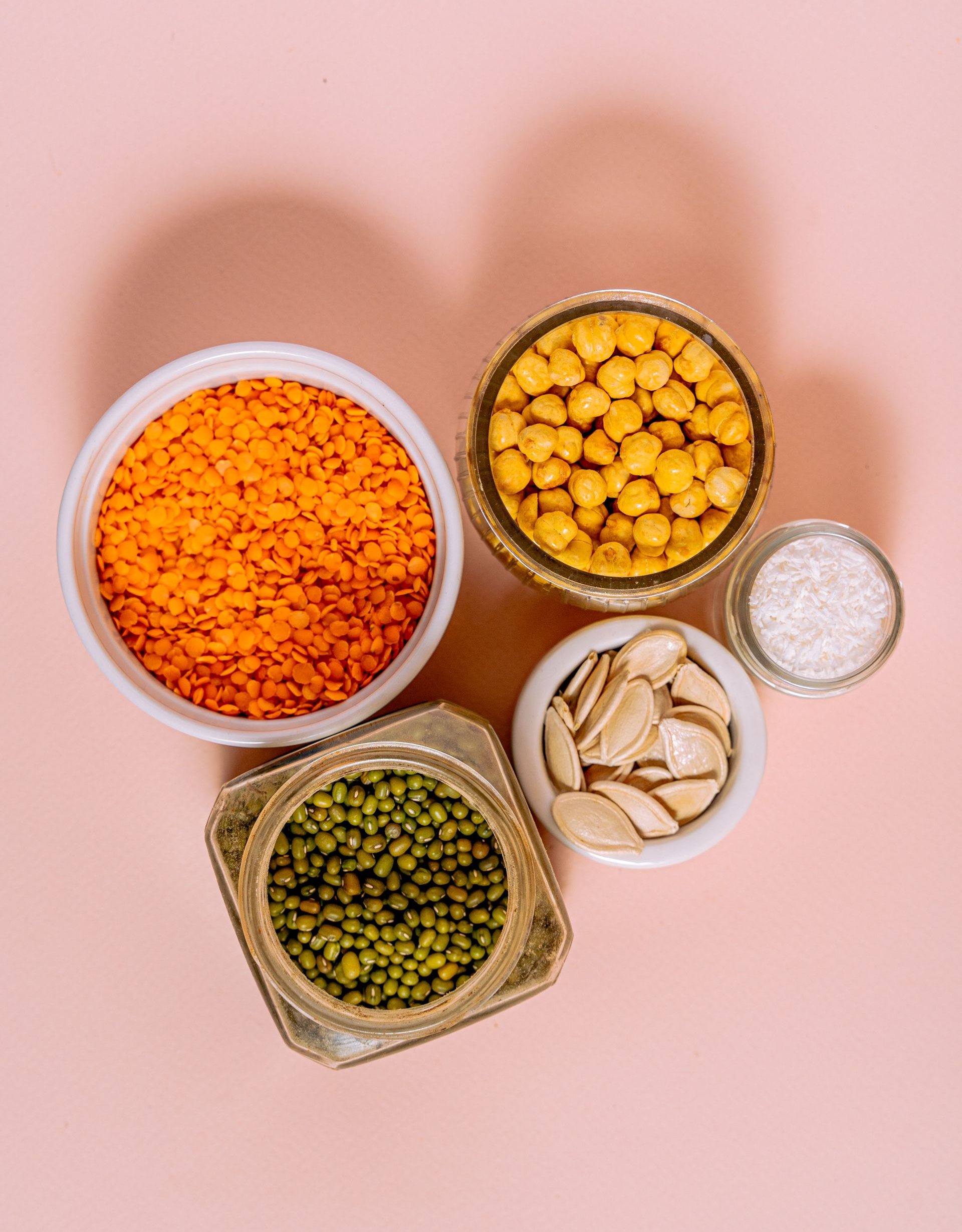 Assorted colorful dry lentils and grains in small bowls on a pink background