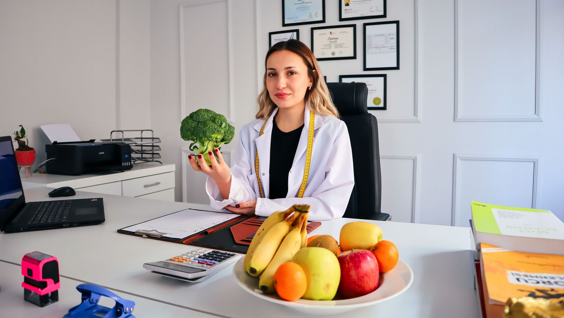 Woman at a desk holding broccoli in an office, with fruit bowl and papers nearby