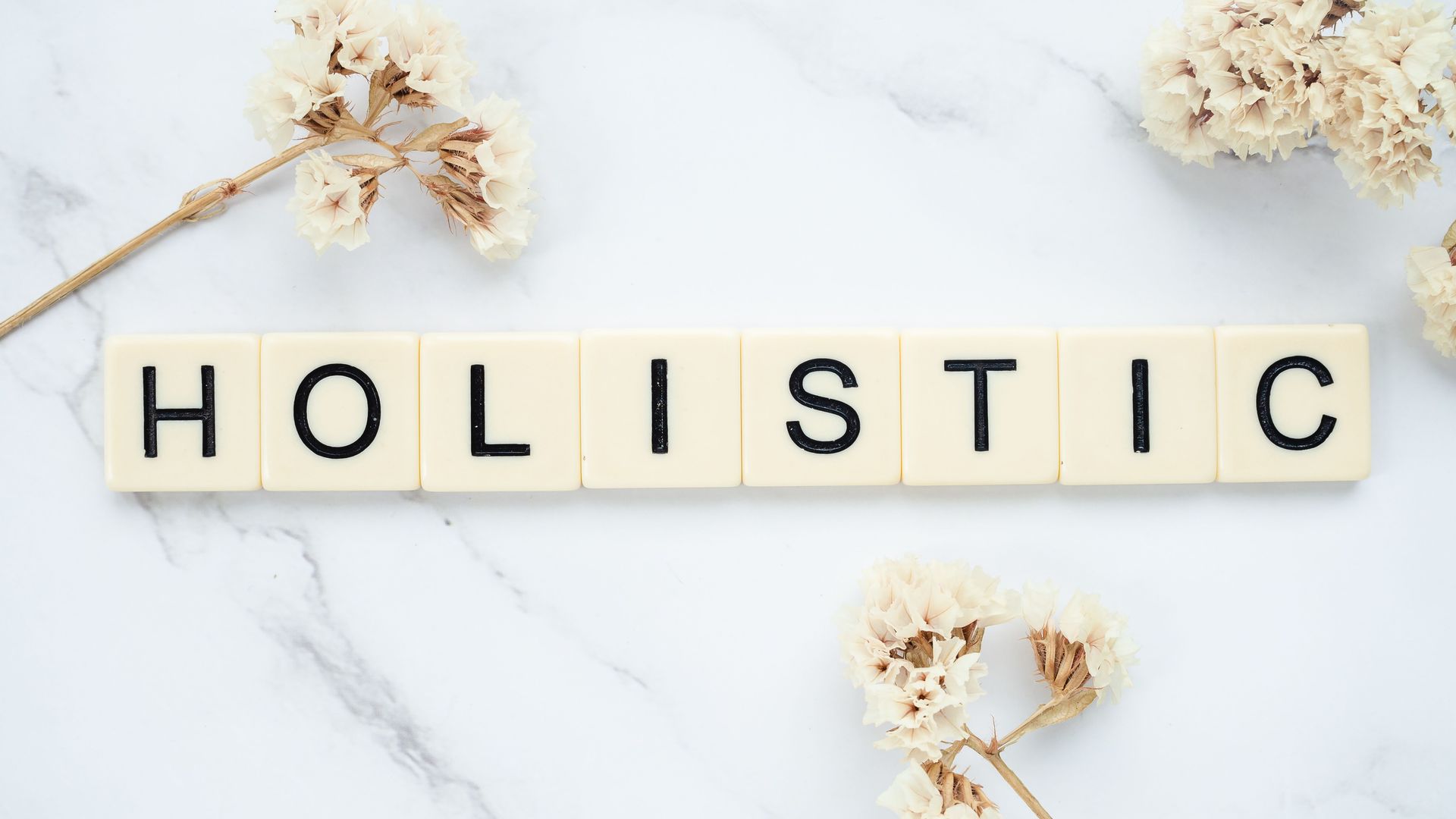H O L I S T I C spelled in wooden letter tiles on a white marble background with dried flowers