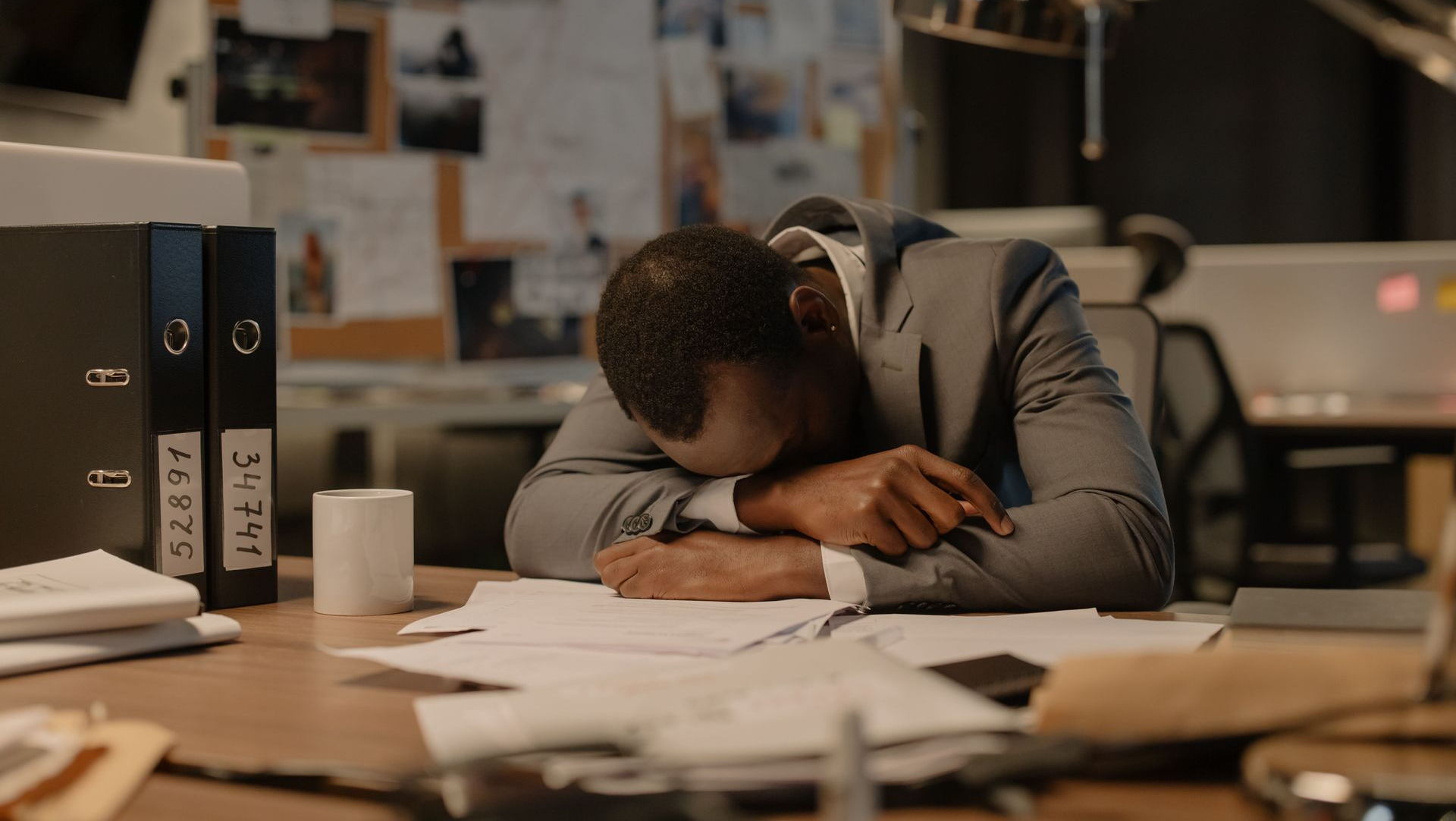 Tired office worker asleep at a desk, head on folded arms amid papers and binders