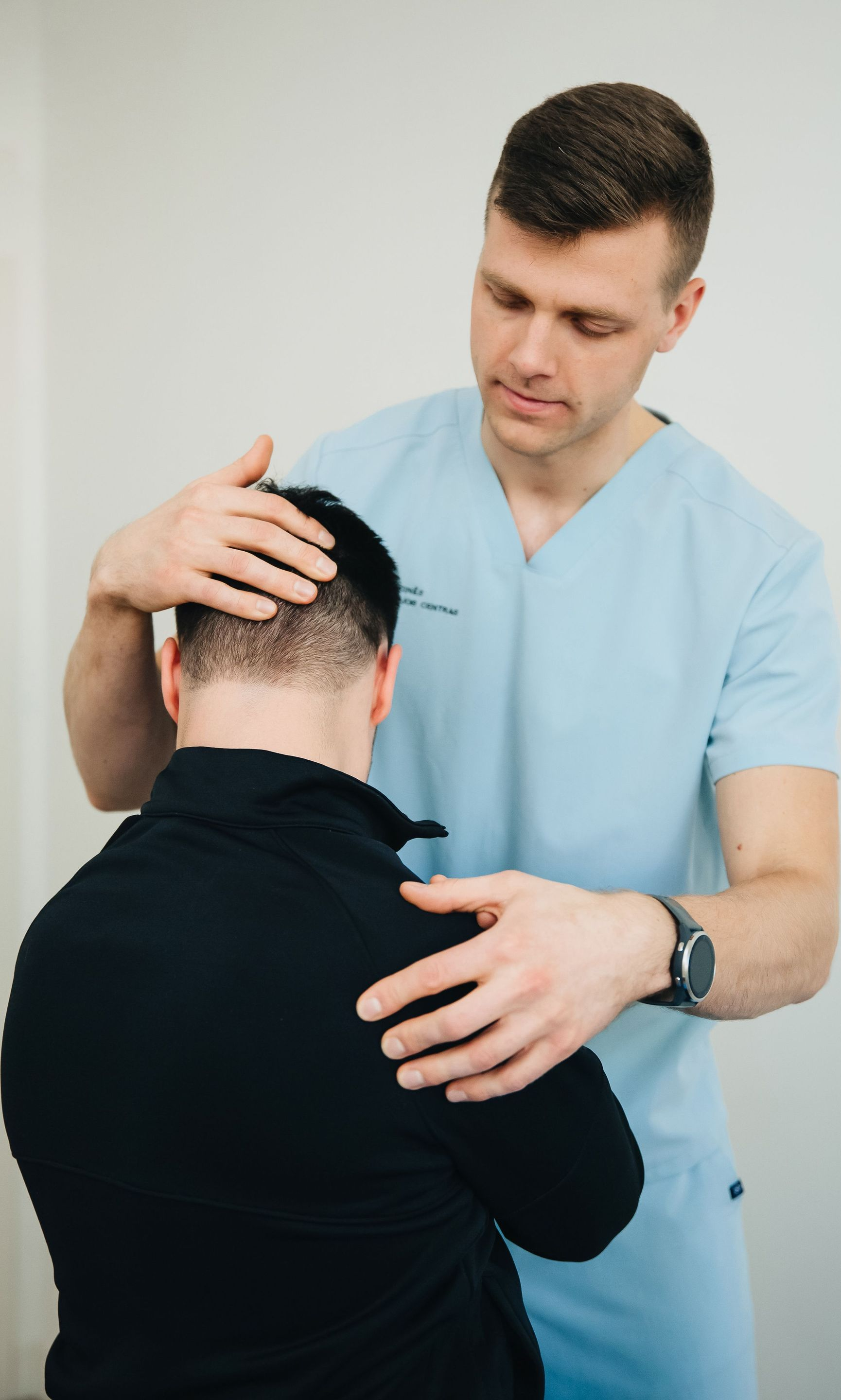 Medical worker in light blue scrubs examining a seated patient’s neck and shoulder.