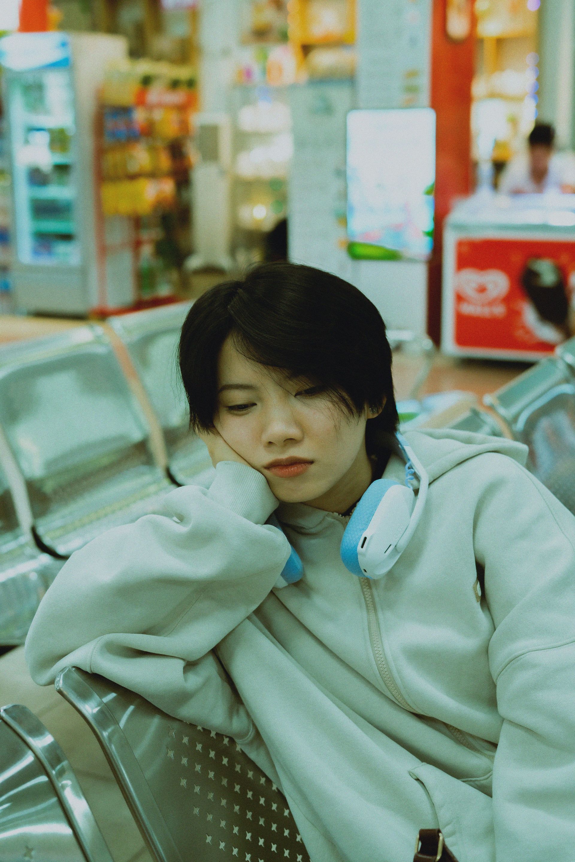 Person resting on a metal bench in a store, wearing a gray hoodie and blue headphones.
