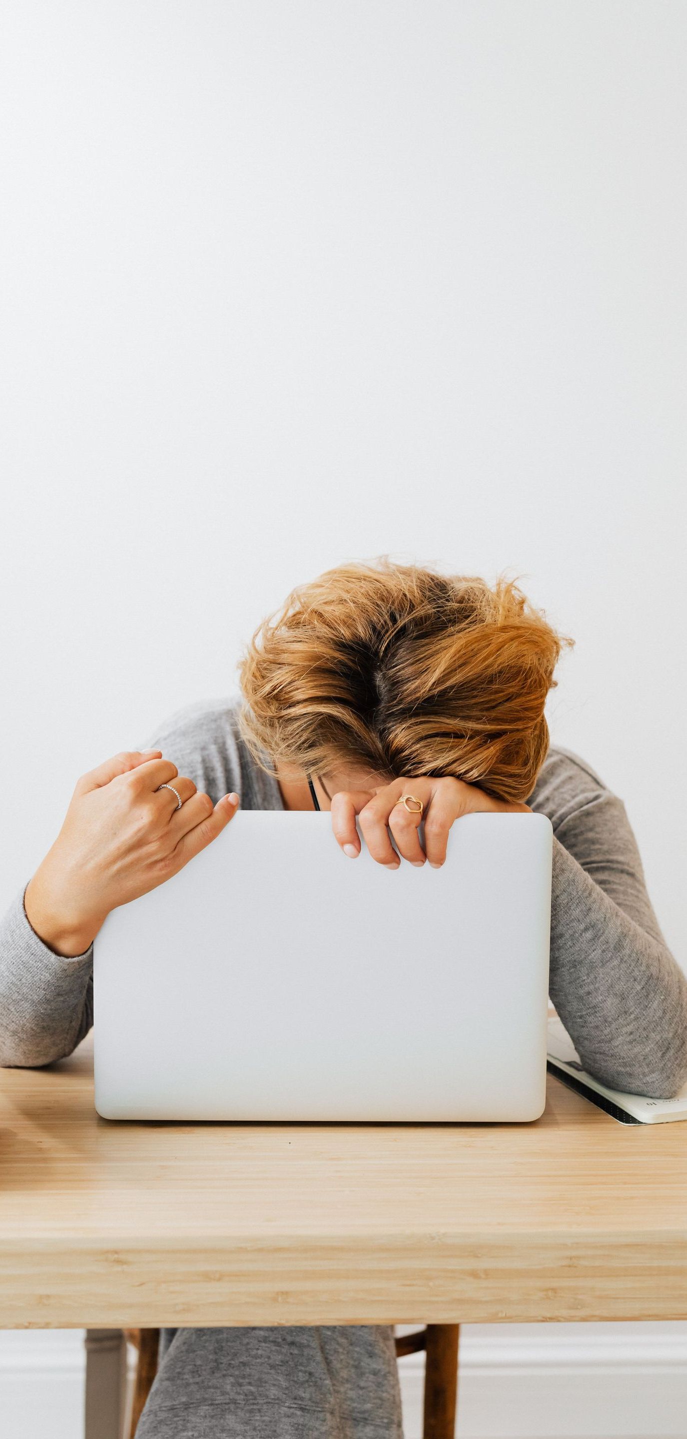 Person resting head on a white laptop at a wooden table against a plain light background