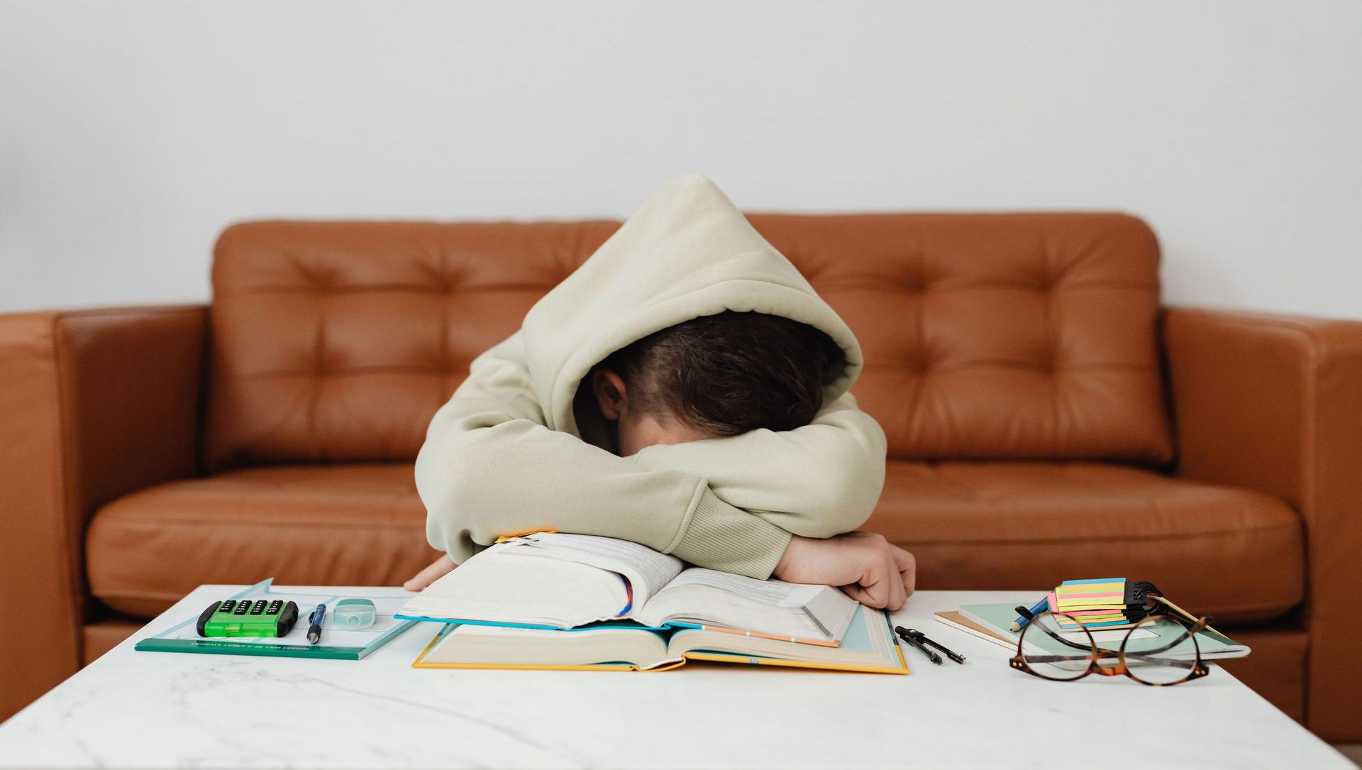 Person sleeping face-down over an open book at a white table, with a brown couch behind.