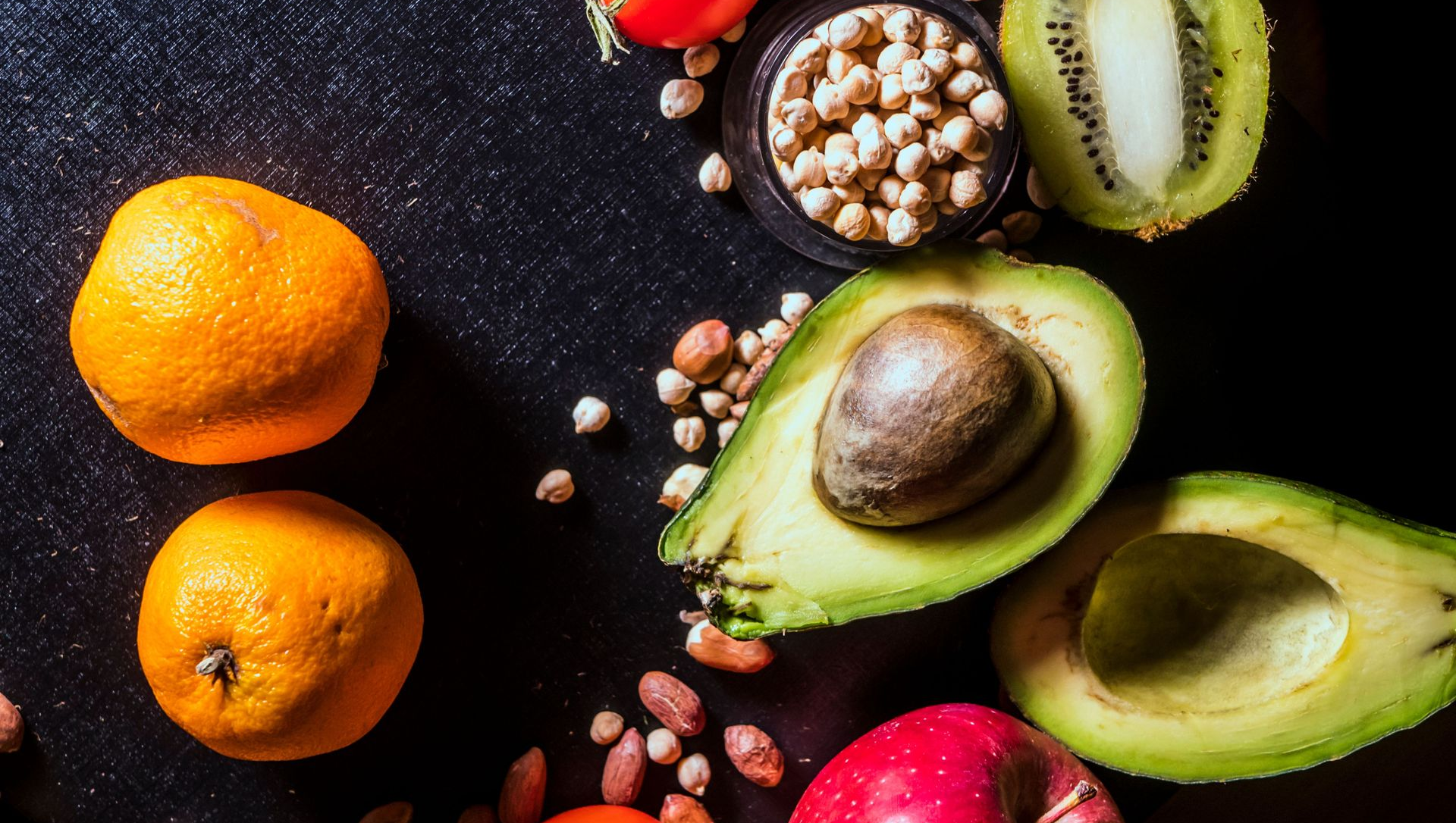 Assorted fruit on a dark surface, including avocados, oranges, kiwi, nuts, and a pomegranate.