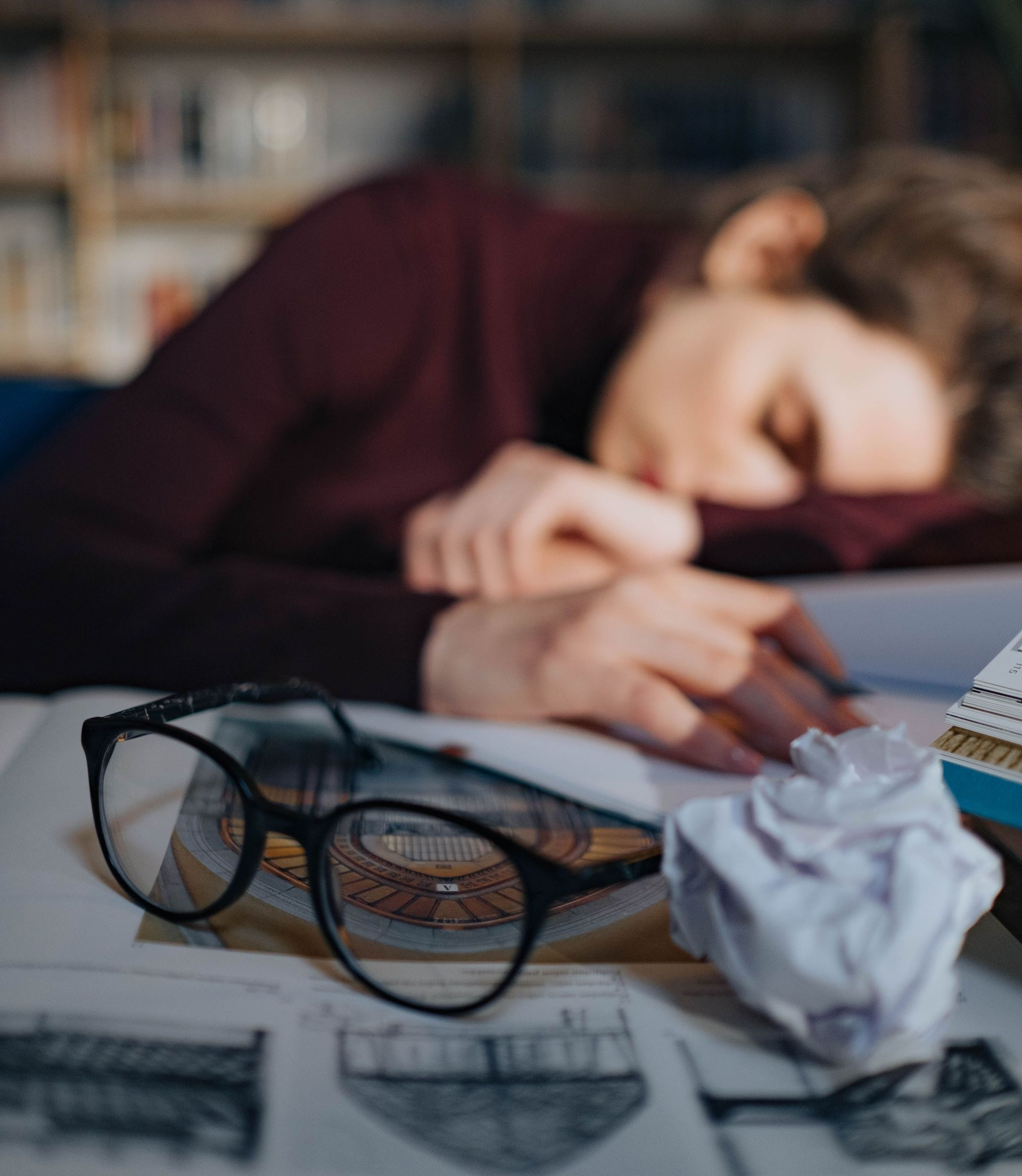Blurry person asleep at a desk beside eyeglasses, crumpled paper, and sketches on paper