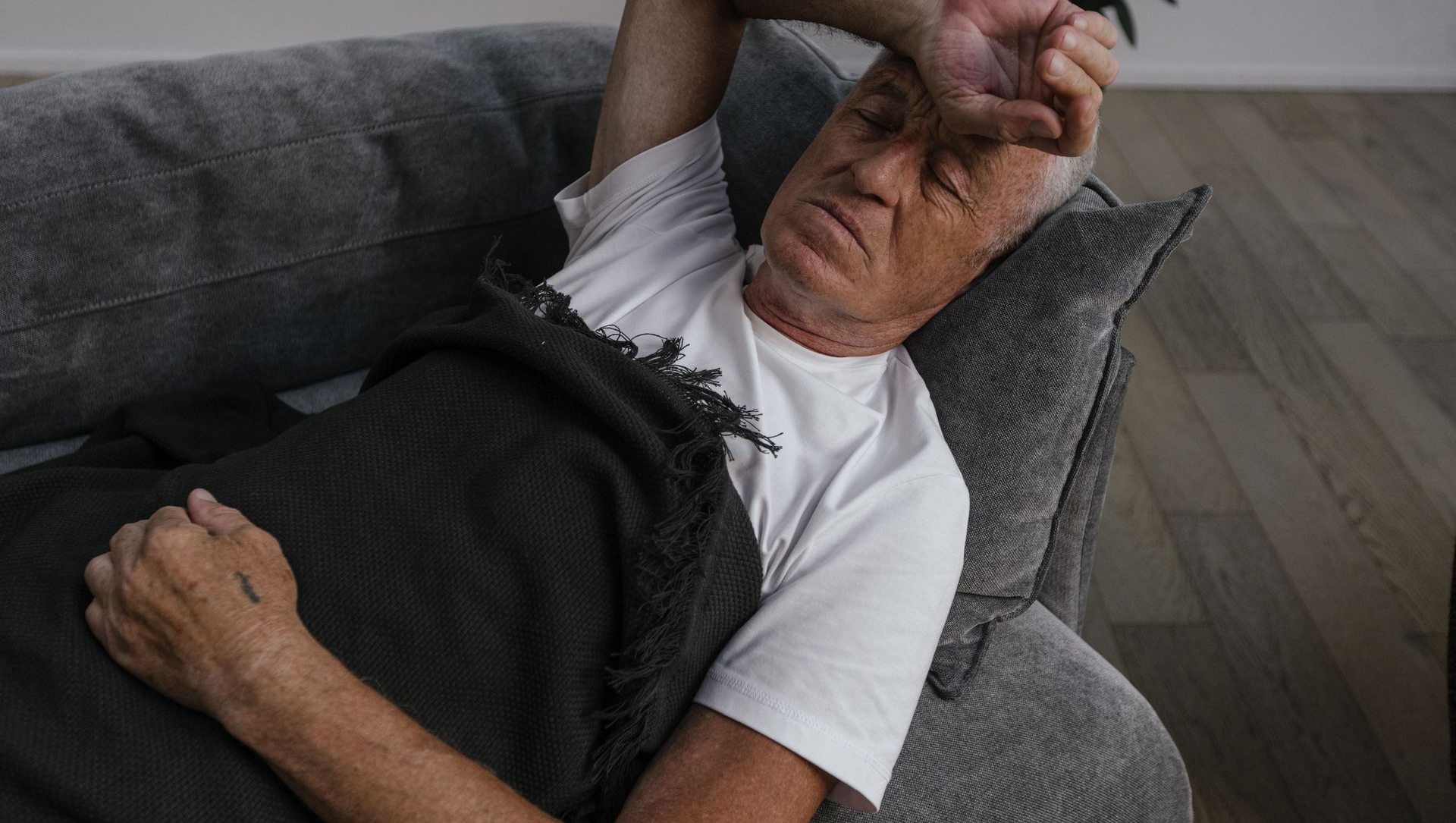 Man lying on a couch, covering his face, appearing tired or unwell under a dark blanket.