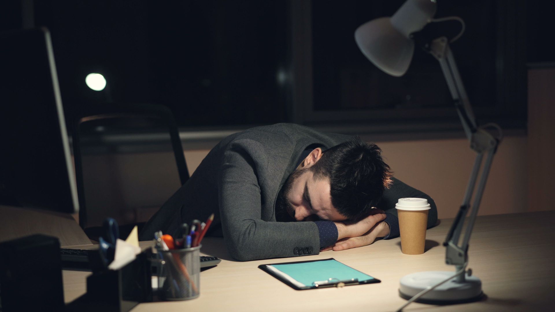 A person sleeps at a desk under a lamp, with a laptop, coffee cup, and tablet nearby.
