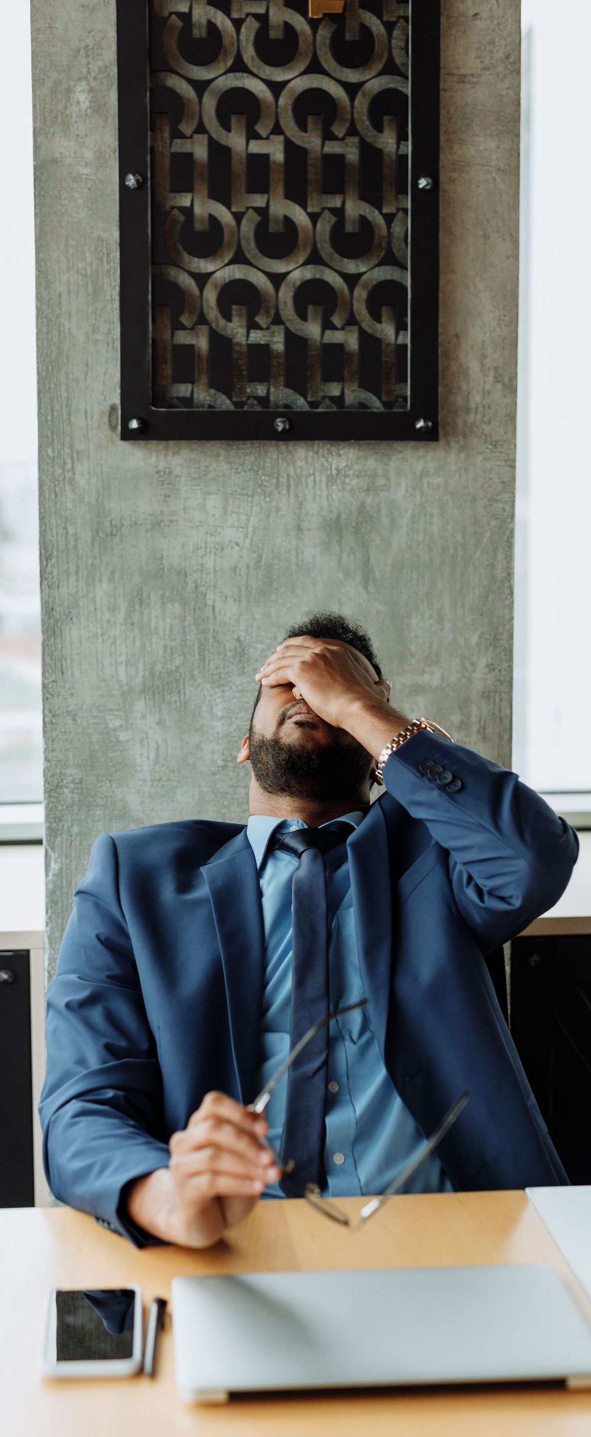 Man in a blue shirt sitting at a desk, rubbing his forehead in apparent stress with a laptop in front of him
