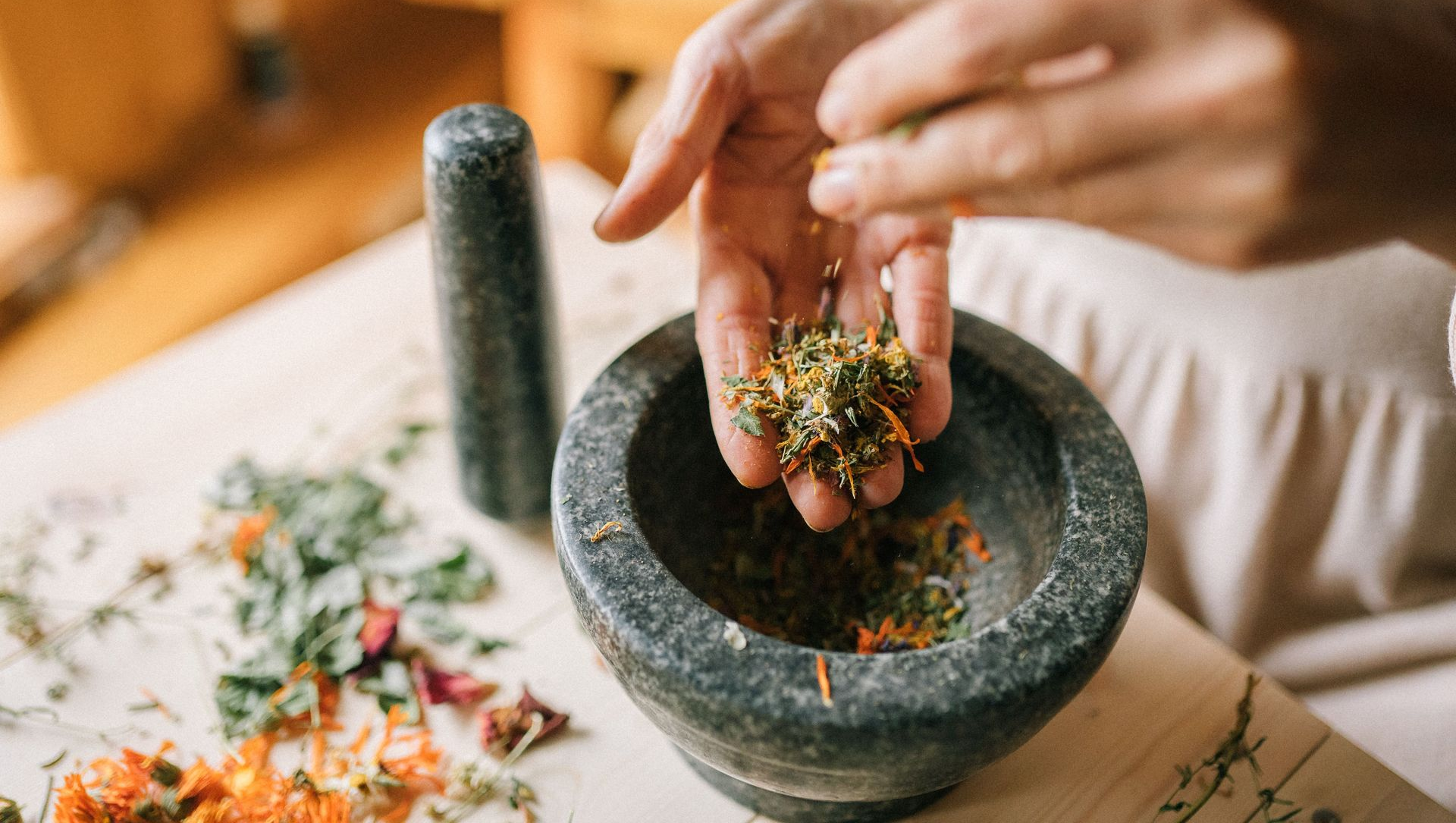 Hand grinding herbs in a stone mortar and pestle on a table with scattered dried flowers