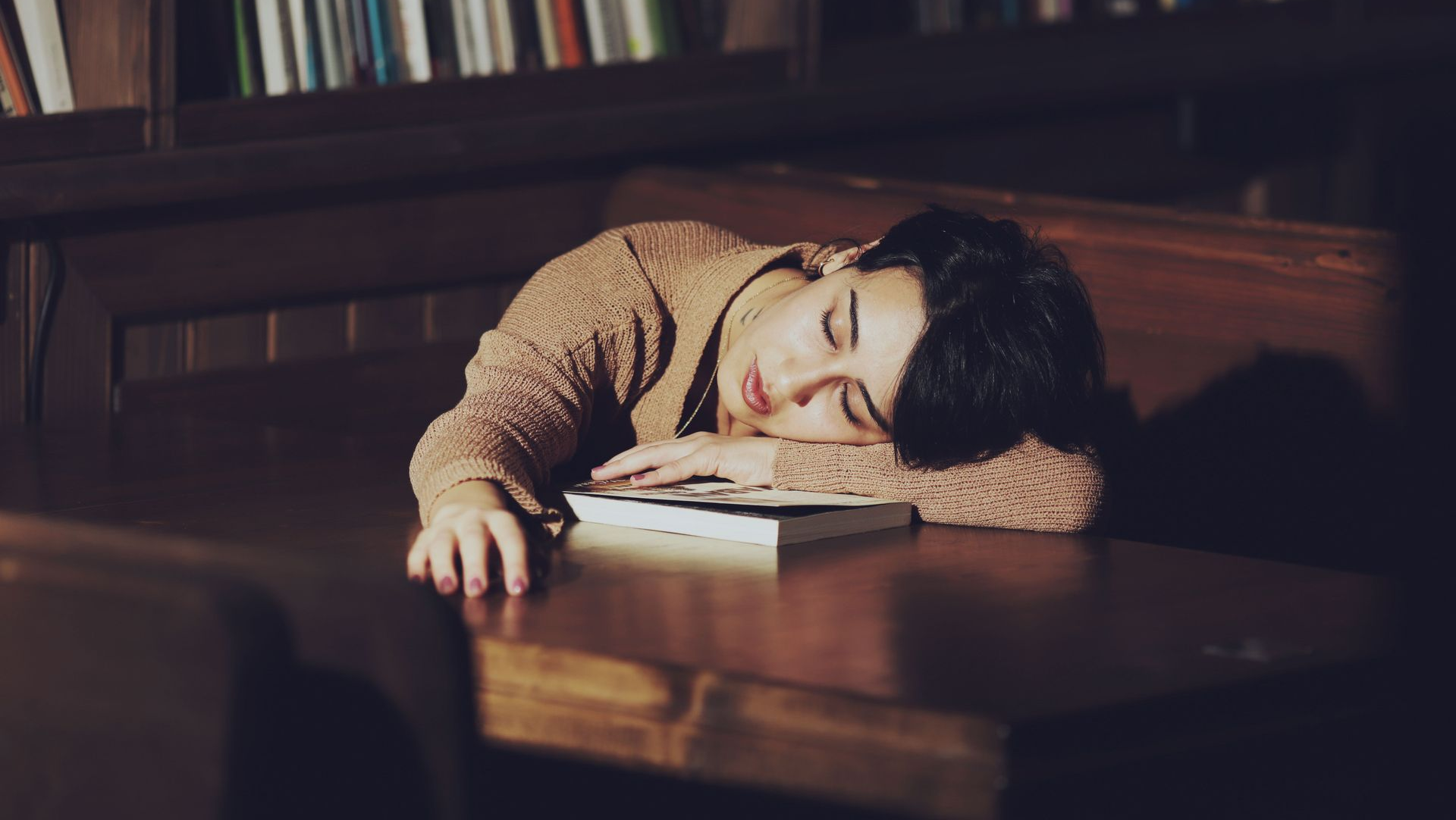Person asleep at a wooden table with head on arms, in a dim library or study.