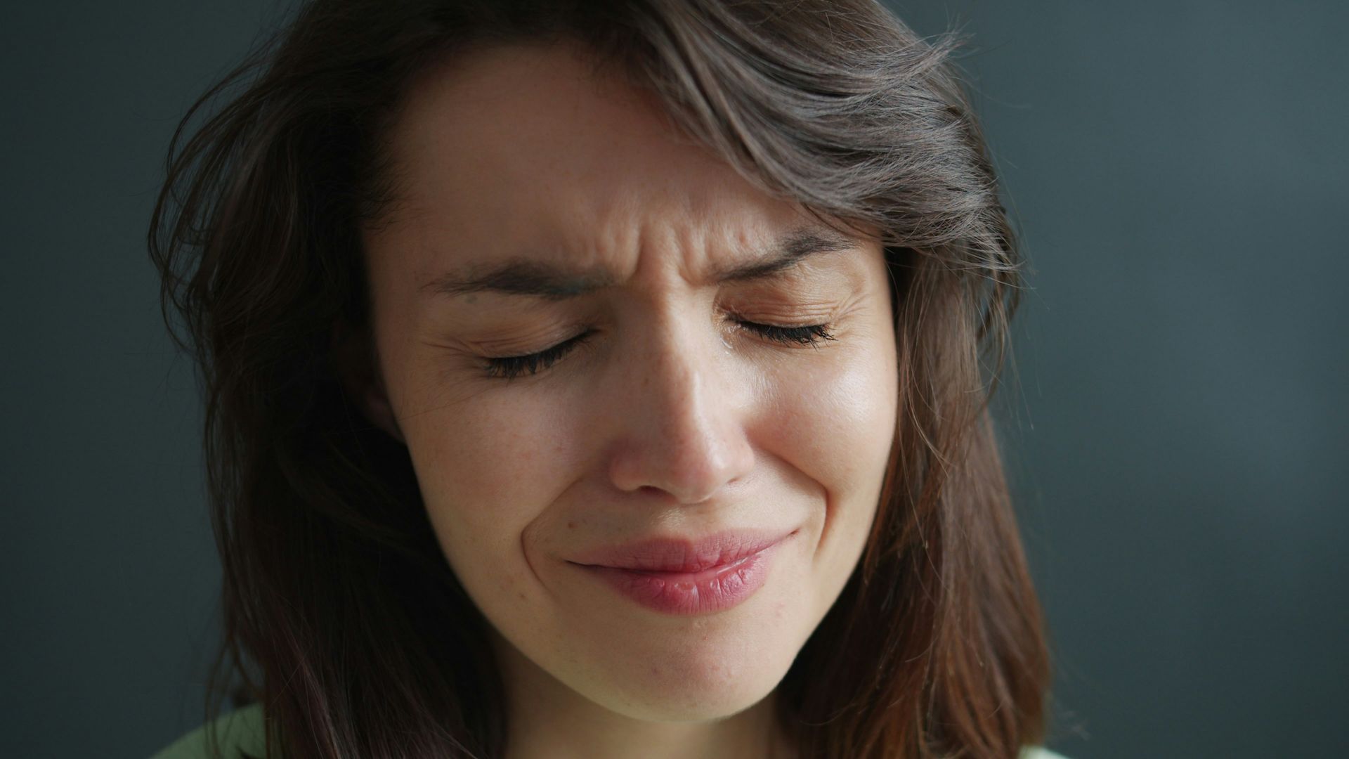 Close-up of a person with eyes closed and a pained expression, against a dark background