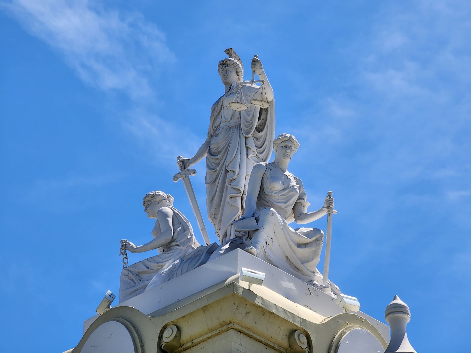 A white stone sculpture of three figures atop a building against a bright blue sky, holding a scale and swords.