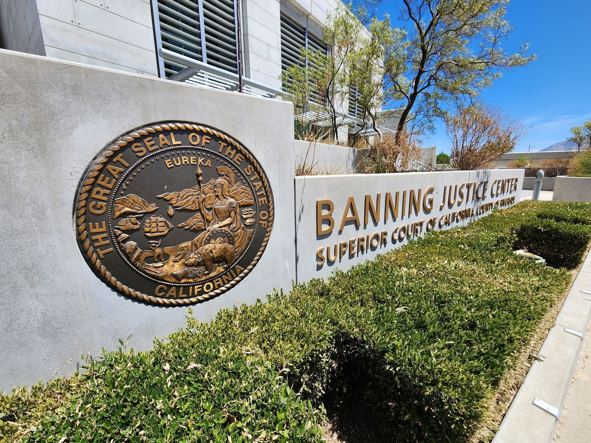 A bronze California state seal mounted on a concrete wall at the Banning Justice Center, fronted by green bushes.