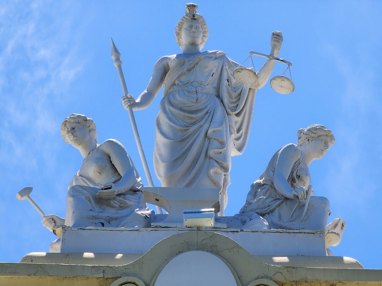 A white sculptural group featuring a central figure holding a staff and scales, flanked by two figures against a blue sky.