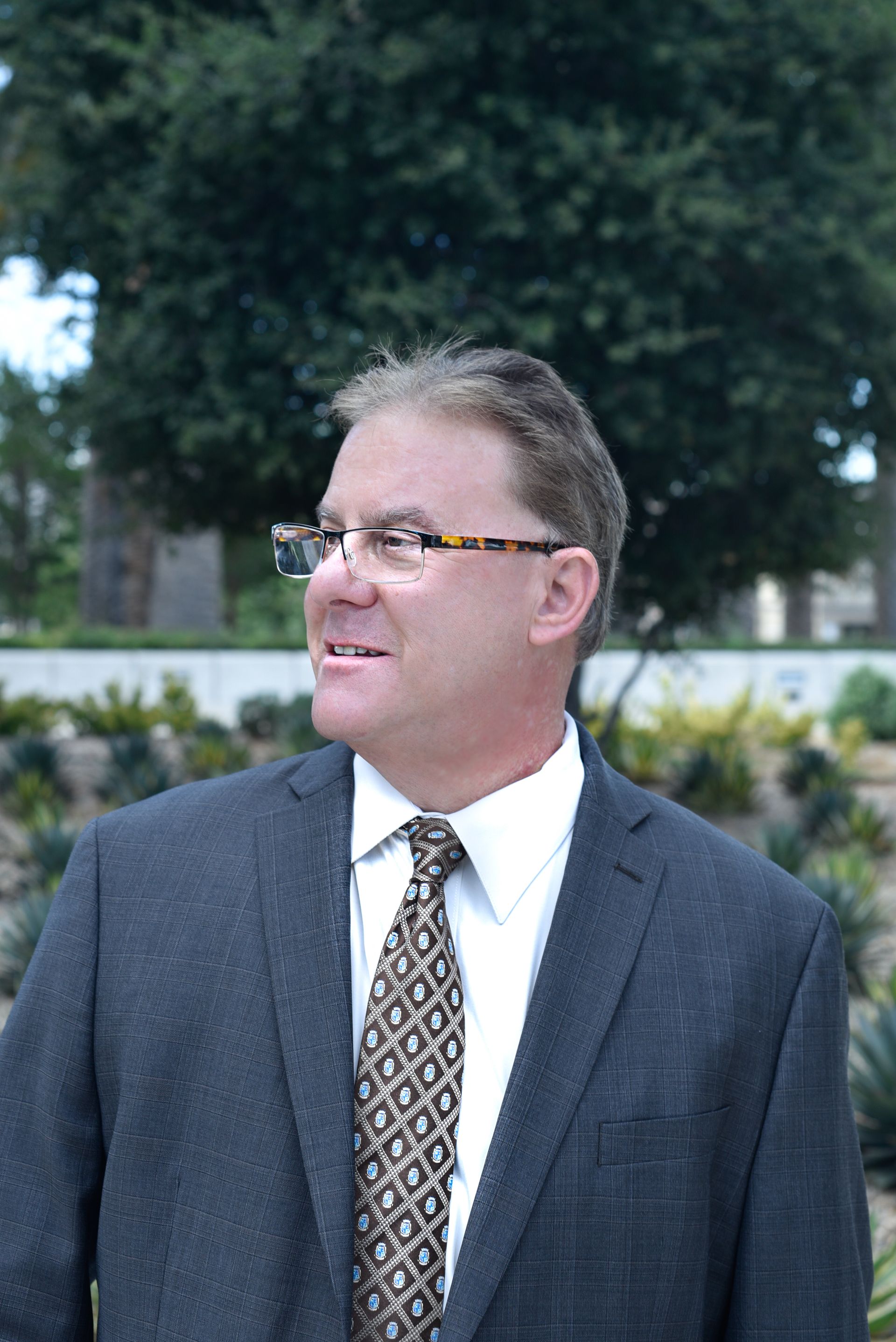 A person with glasses, wearing a dark blazer and a patterned tie, stands in a garden and looks to the left.